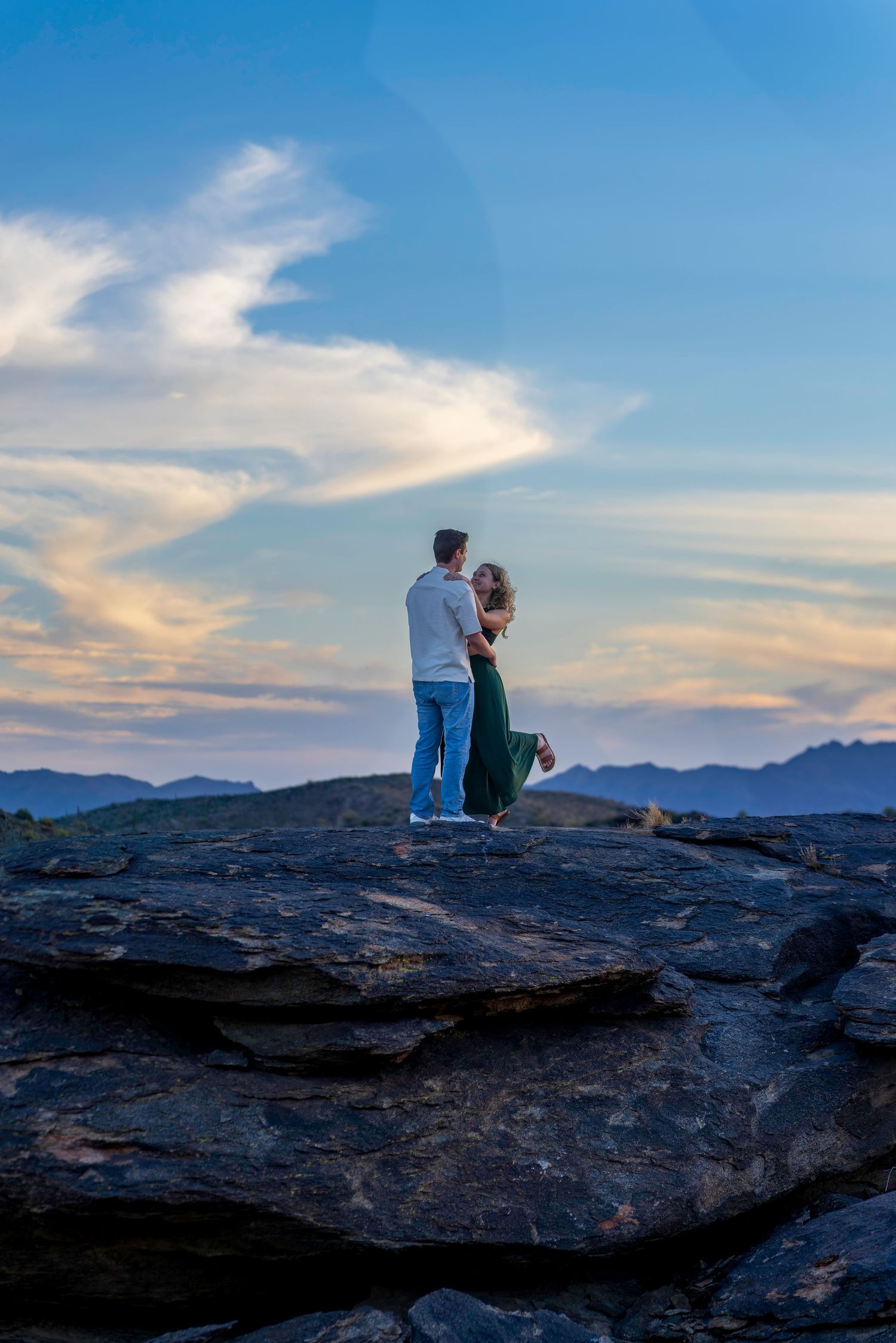 Couple embracing on rocky hilltop silhouetted against a blue twilight sky and mountains.