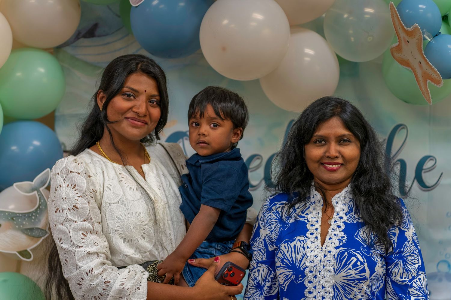 Two Indian women and a toddler boy pose in front of a balloon arch at a birthday party.