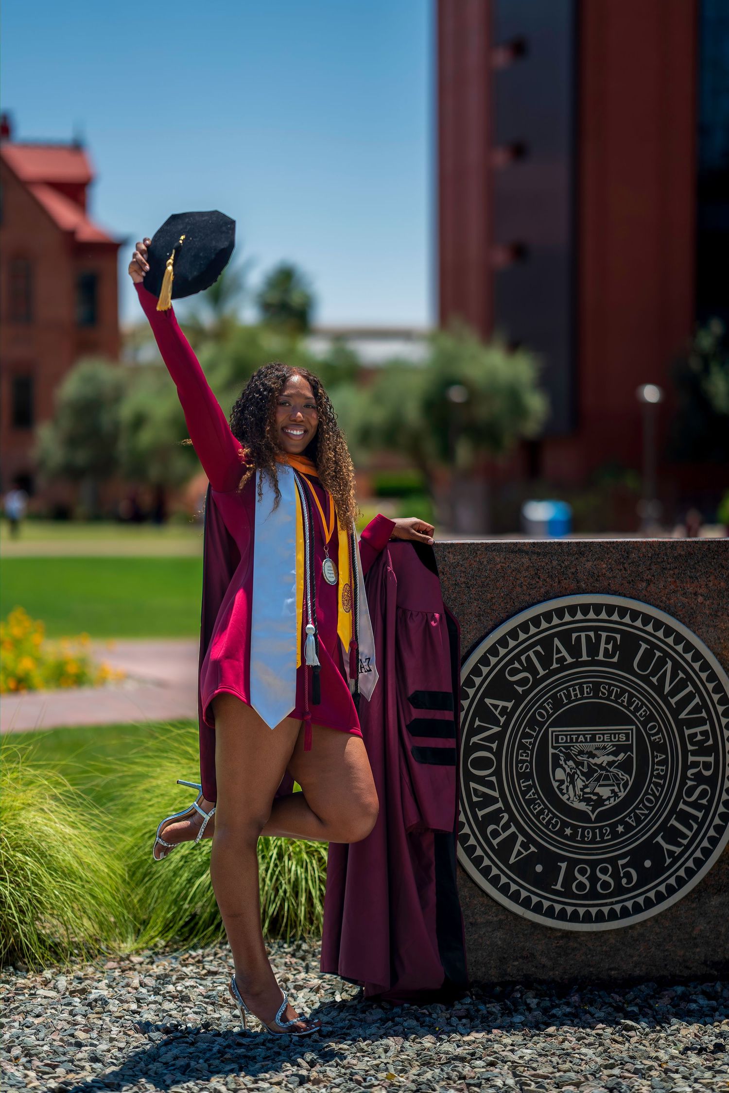 Happy graduate raising cap beside Arizona State University 1885 seal in maroon doctoral regalia.