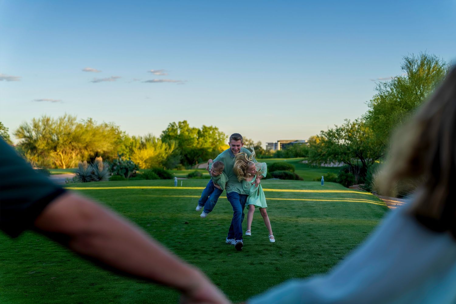 Family playfully reuniting on lush green grass during golden hour outdoor photography session.
