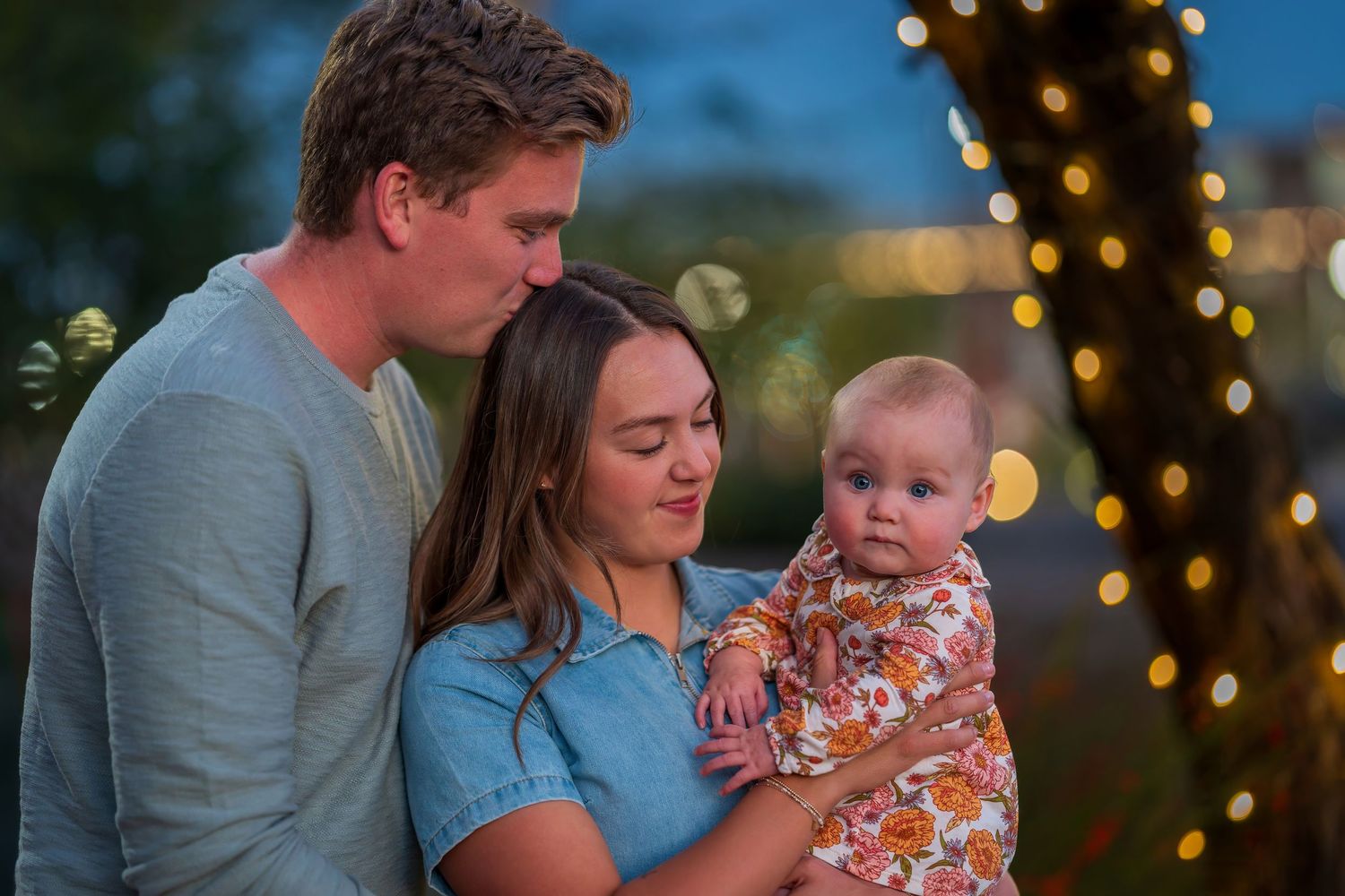 Young couple sharing a tender moment kissing while holding their baby outdoors with bokeh lights in background.