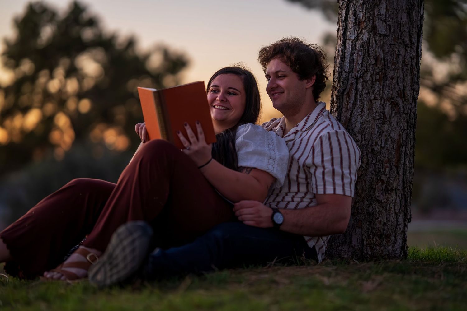 Couple relaxes against tree at sunset, sharing a book on grass in a park.