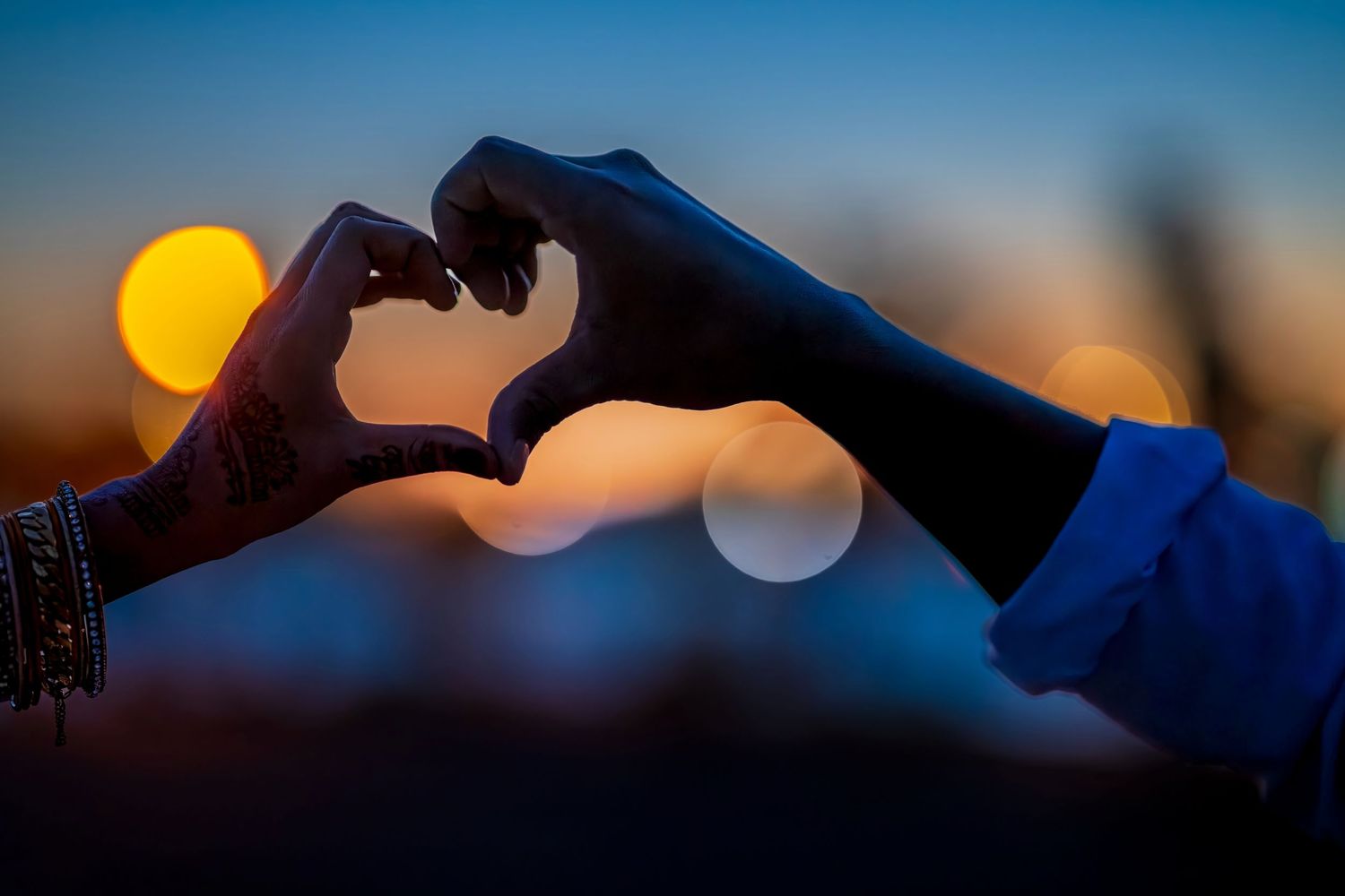 Two hands form a heart shape silhouetted against a colorful bokeh sunset sky.