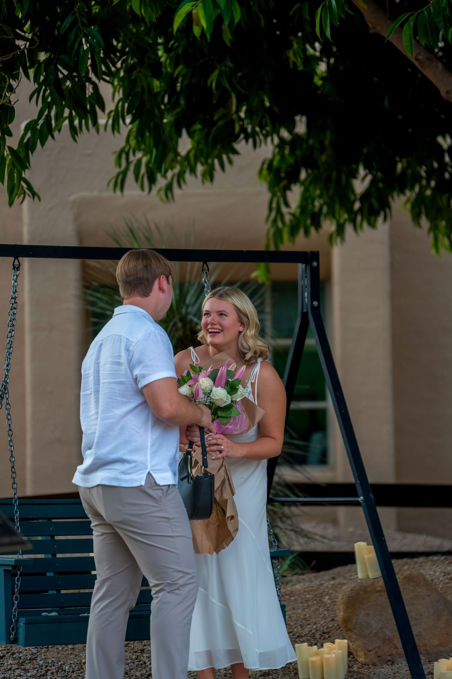 Smiling woman in white dress receives bouquet from man near swing.