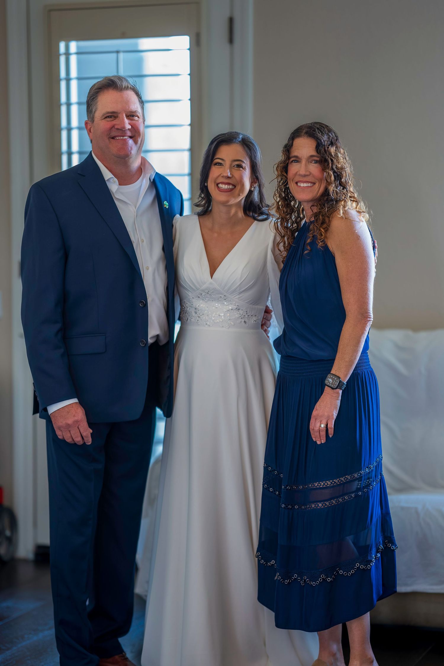 Bride in white gown poses with man in navy suit and woman in blue dress at indoor wedding venue.