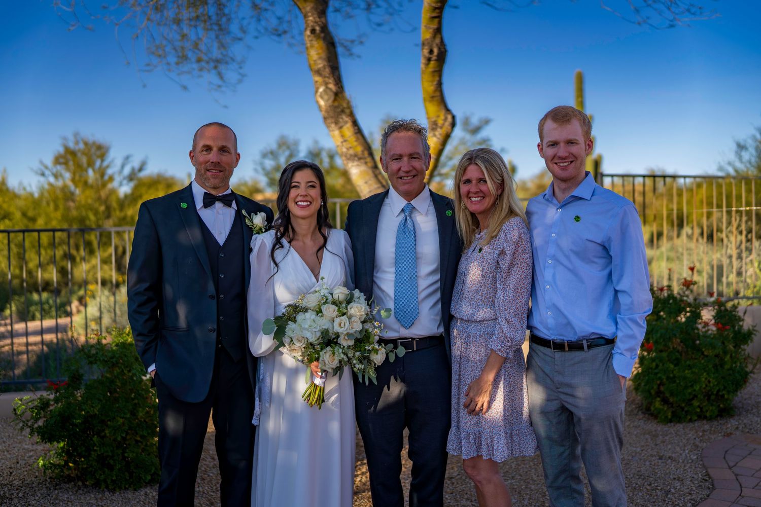 Bride and groom posing with family group of five outdoors under blue sky at wedding.