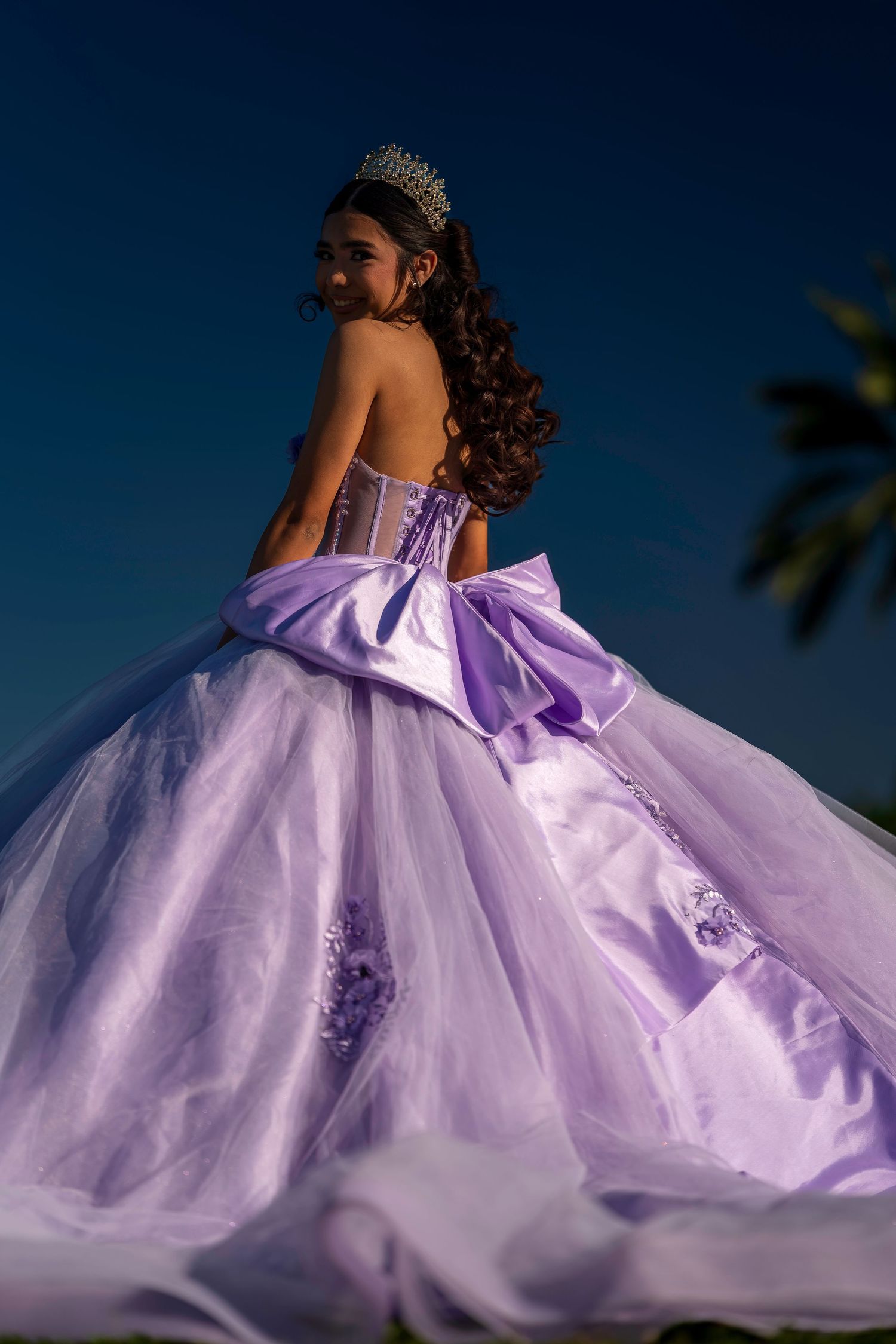 Young Latina girl in lavender quinceañera ball gown with large bow and tiara poses outdoors at dusk.