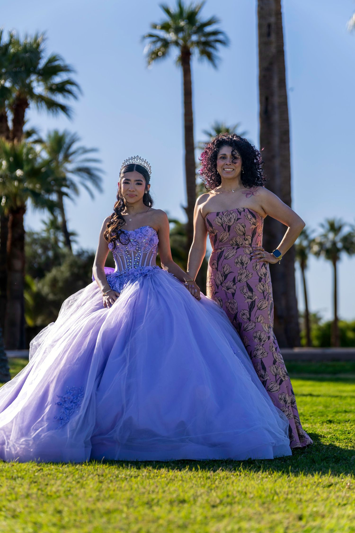Two women in elegant formal gowns pose outdoors among palm trees, one wearing a lavender ball gown with a tiara.