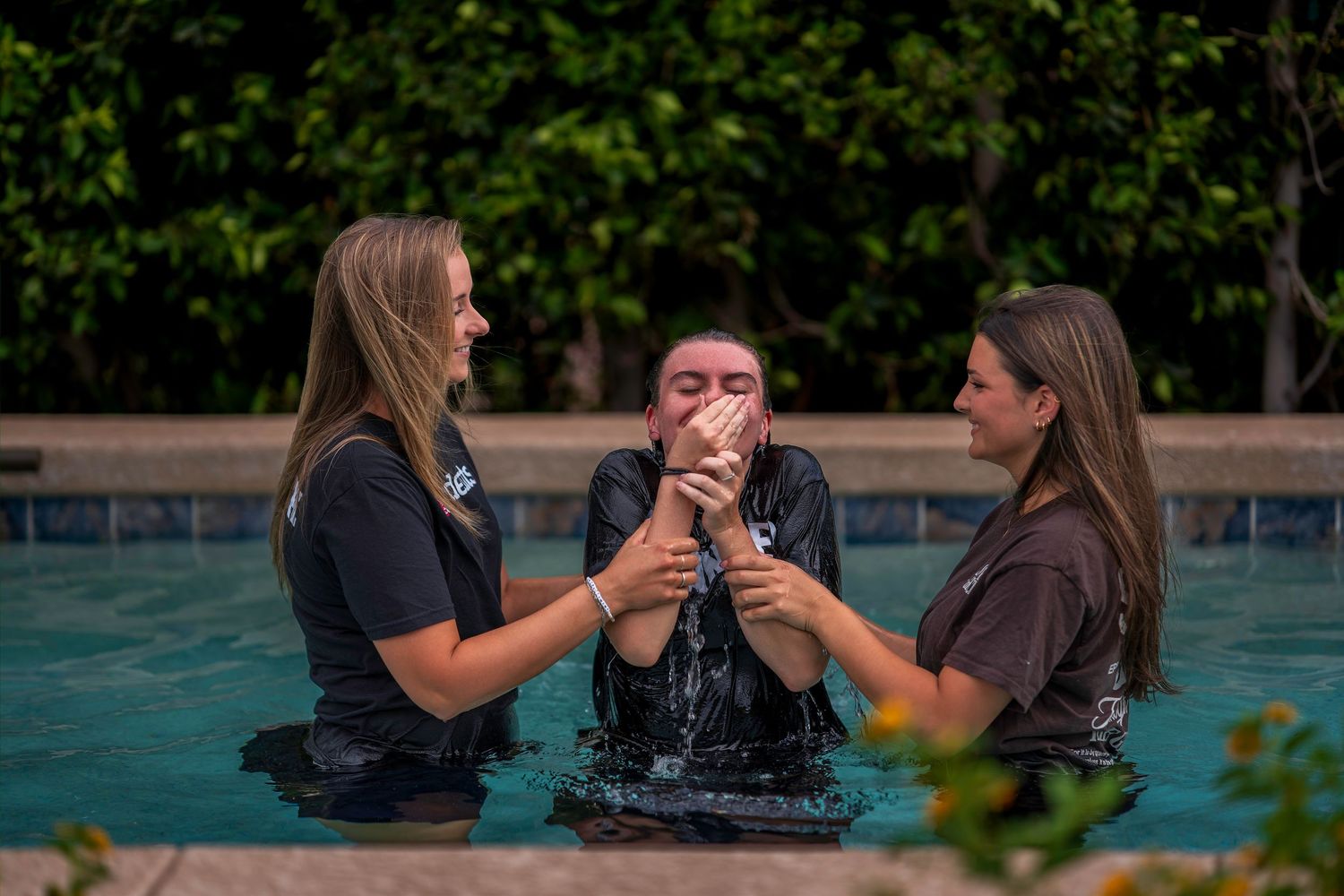 Three people standing in a pool during a baptism ceremony, with two women supporting a person in the center.