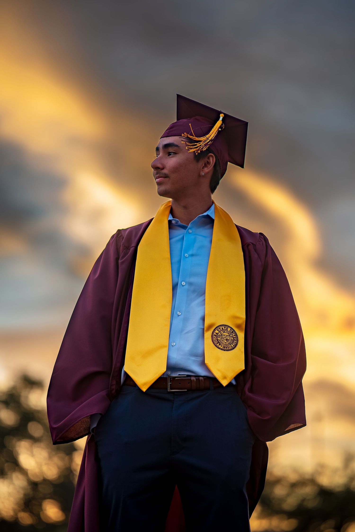 Graduate in maroon and gold regalia photographed against dramatic sunset sky.