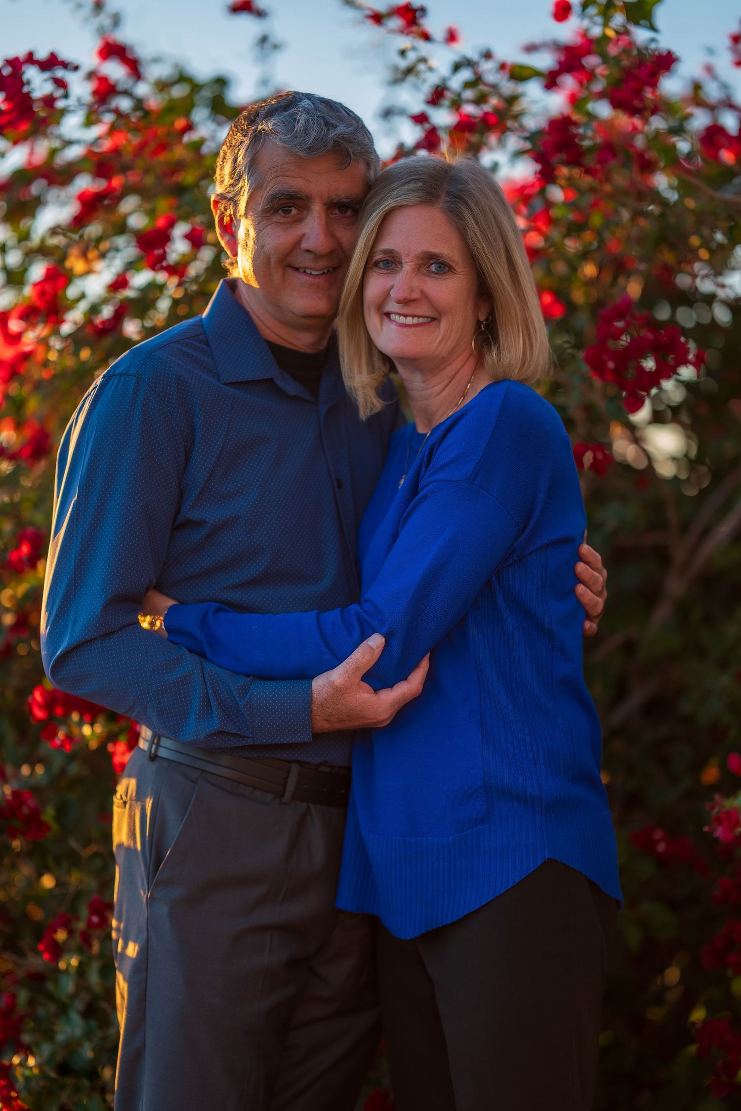 Couple in blue and dark outfits embracing near blooming red bougainvillea bush.