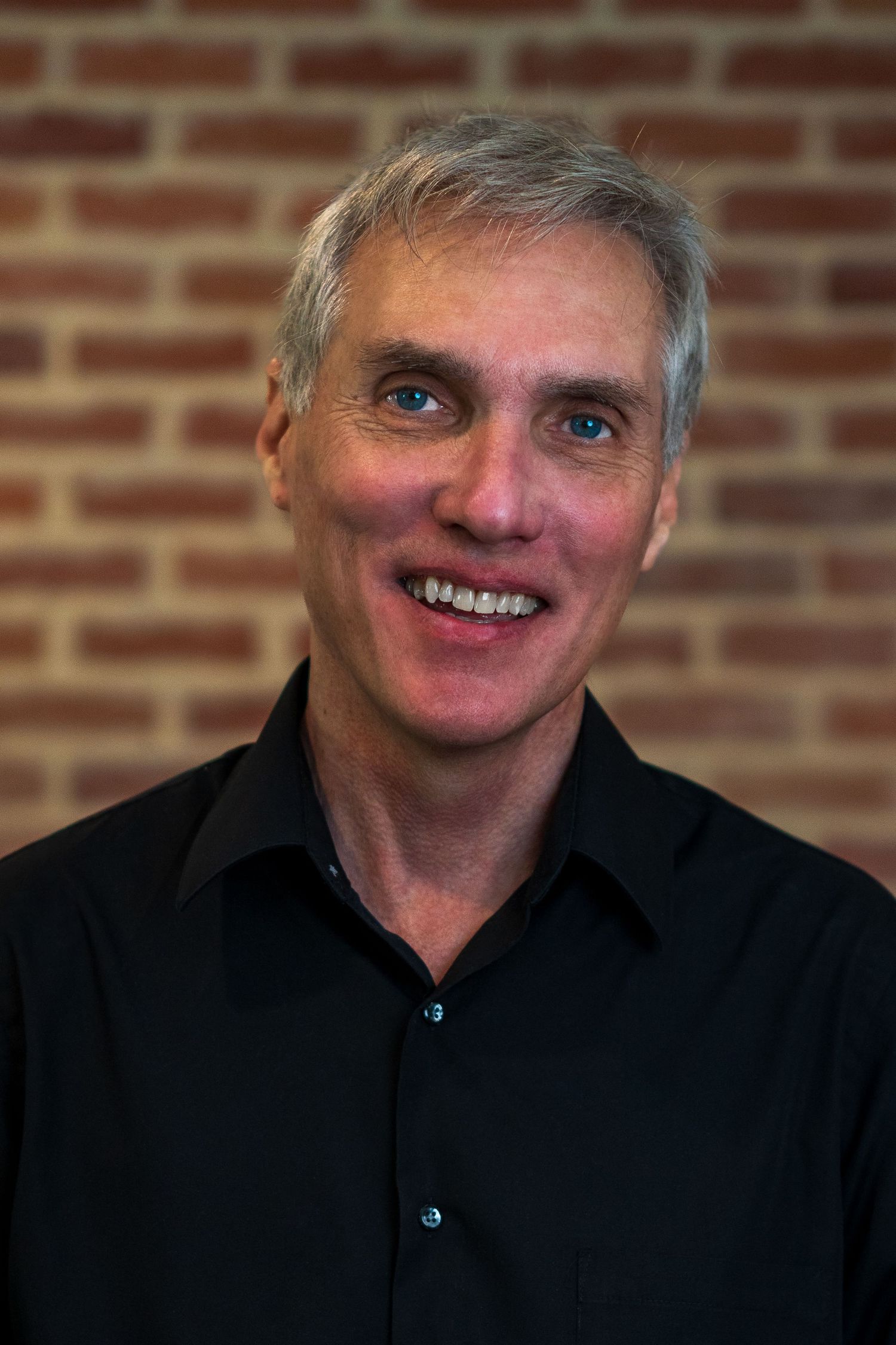 Professional headshot of someone in black attire smiling against a brick wall background.