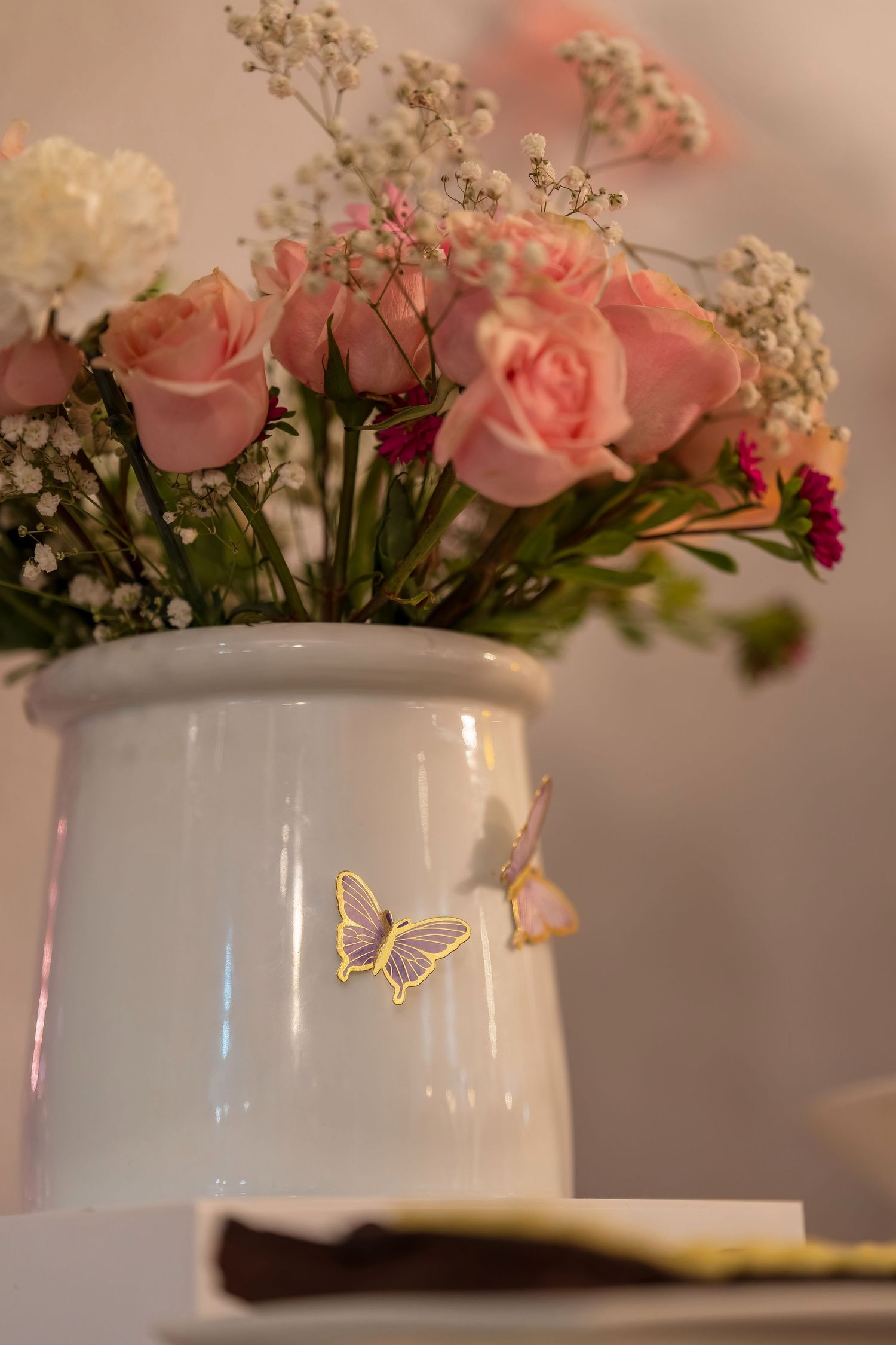 A white vase with pink roses and baby's breath features a delicate golden butterfly decoration.