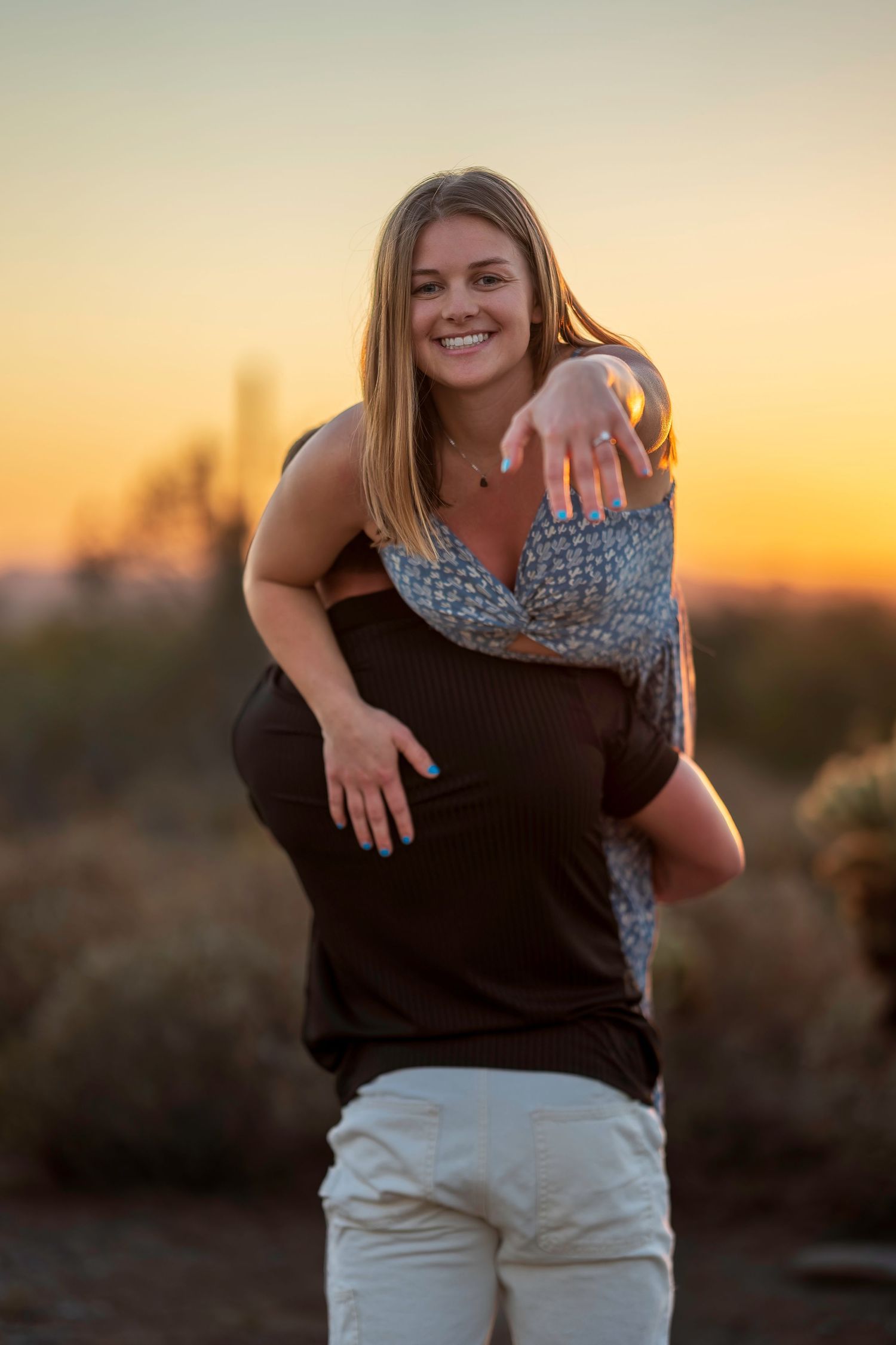 Woman smiling on piggyback ride at desert sunset, wearing blue patterned dress with engagement ring visible.
