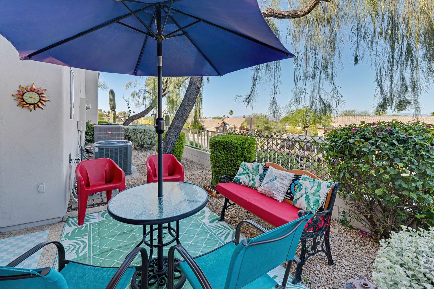 Colorful patio with teal furniture, red chairs, round table, and blue umbrella surrounded by lush greenery.
