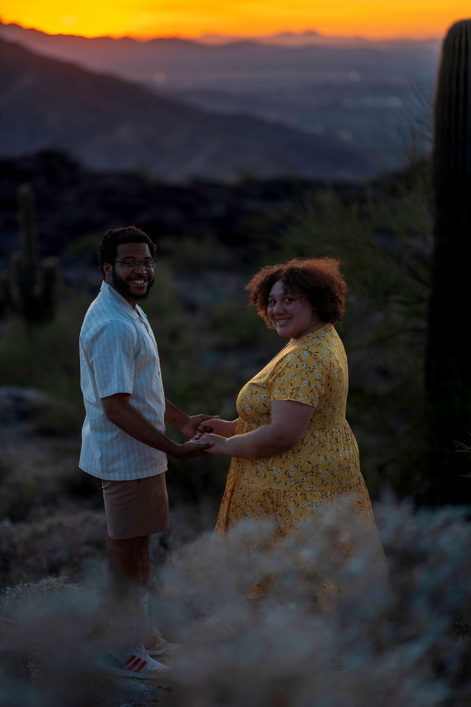 Happy couple holding hands in desert landscape at golden sunset with saguaro cacti in background.