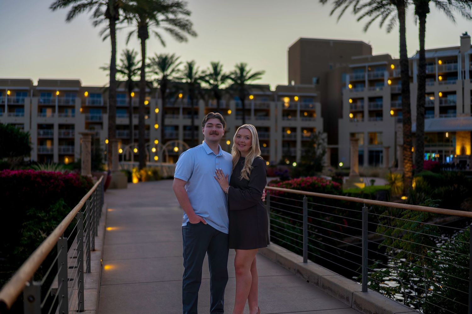 Couple smiling on a bridge at dusk with illuminated resort buildings and palm trees in the background.