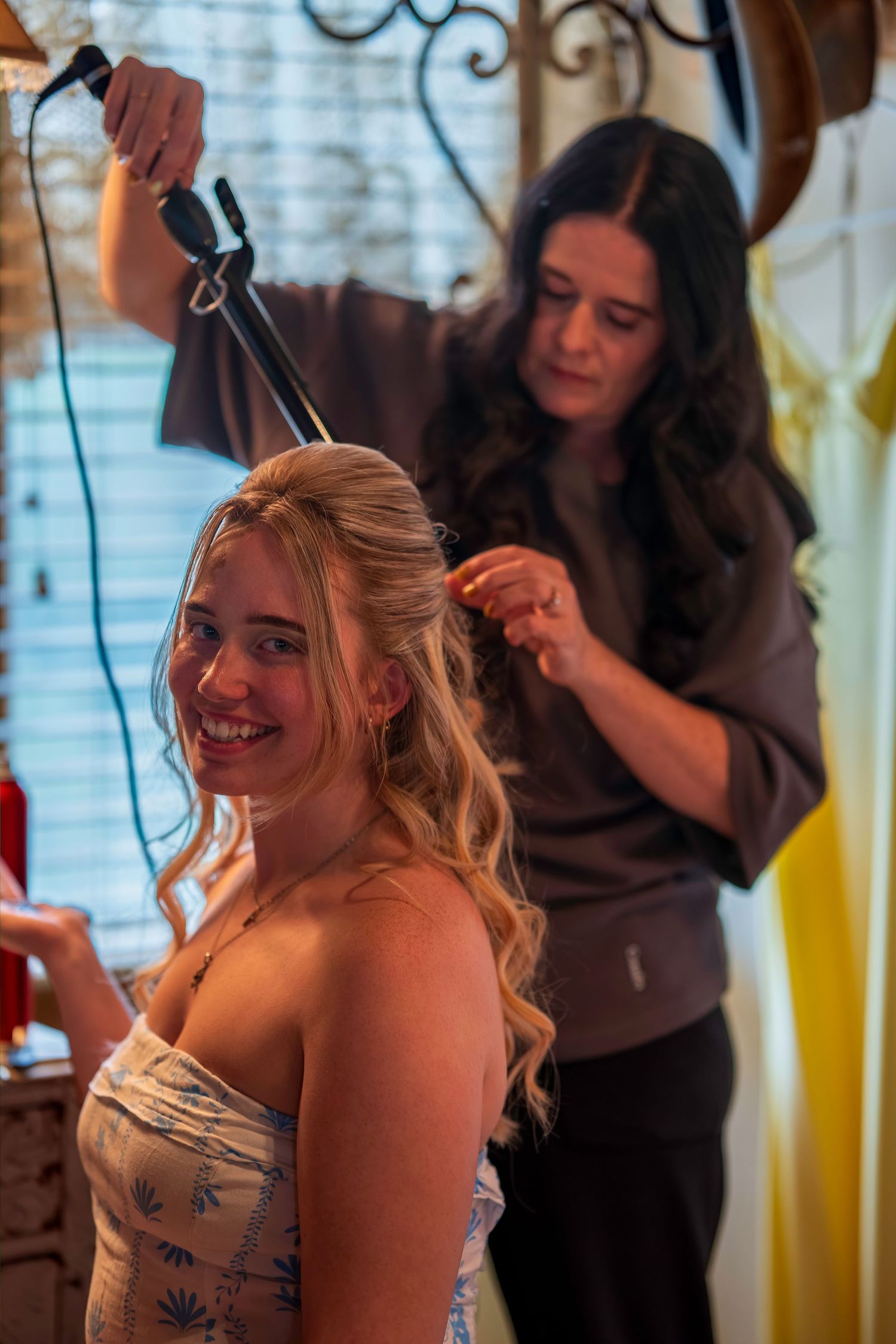 A hairstylist curling the blonde hair of a smiling young woman in a strapless dress during wedding preparations.