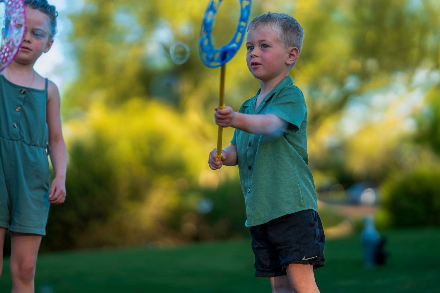 Boy in green shirt plays with bubble wand in sunny park while girl in green dress watches nearby.