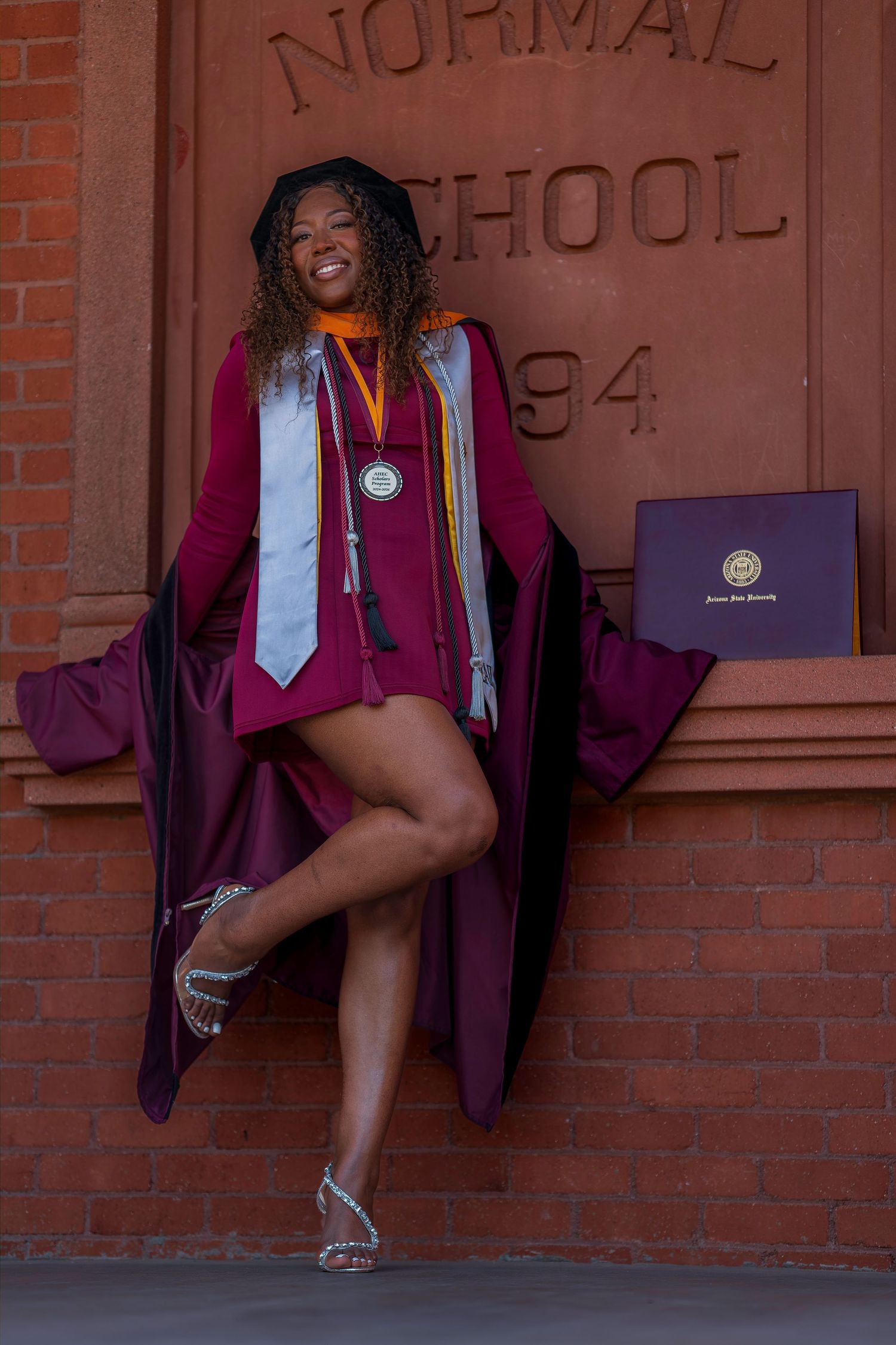 Joyful graduate in maroon cap and gown posing with diploma at ASU Normal School building.