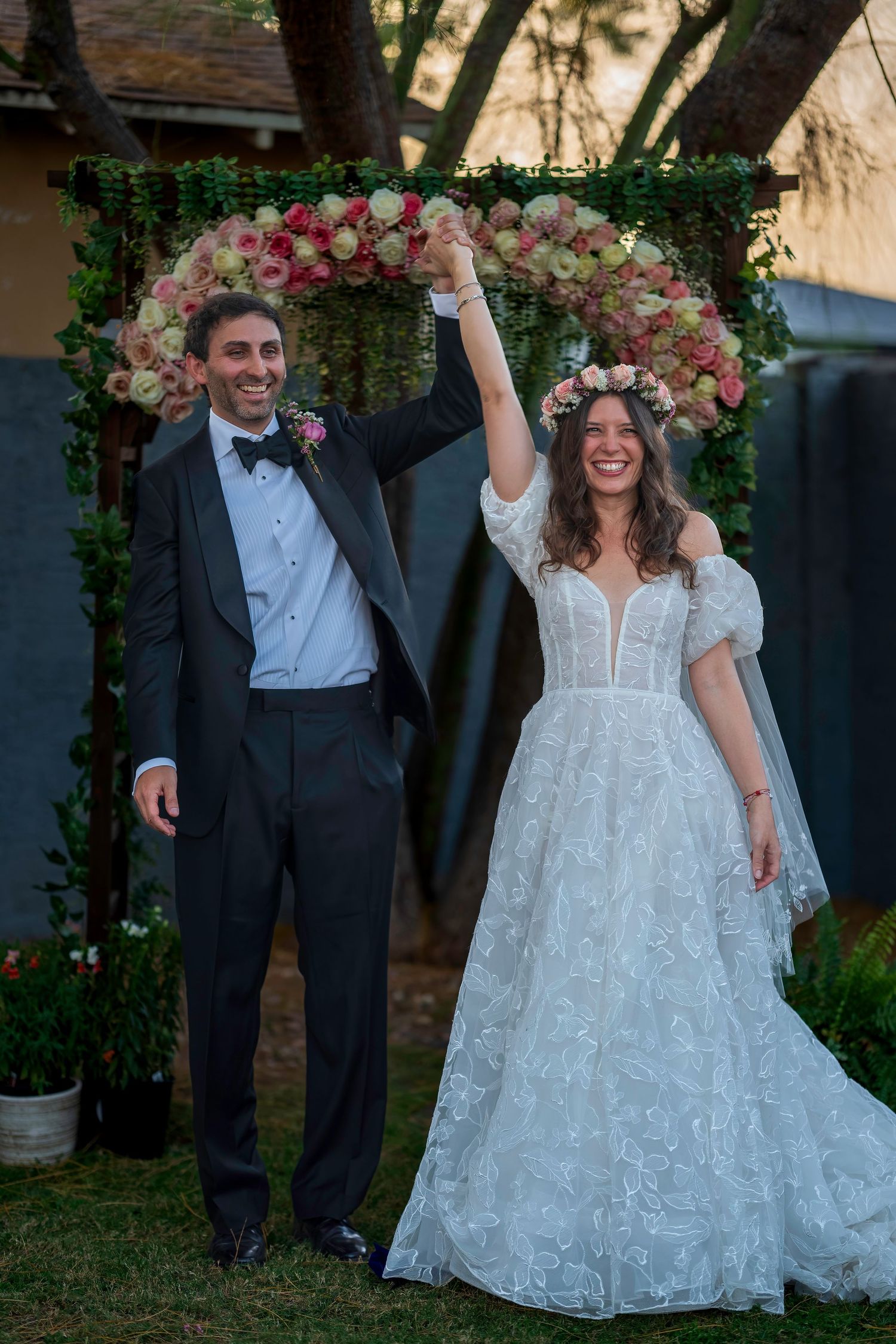 Joyful newlyweds raising hands under floral arch at outdoor garden wedding ceremony.