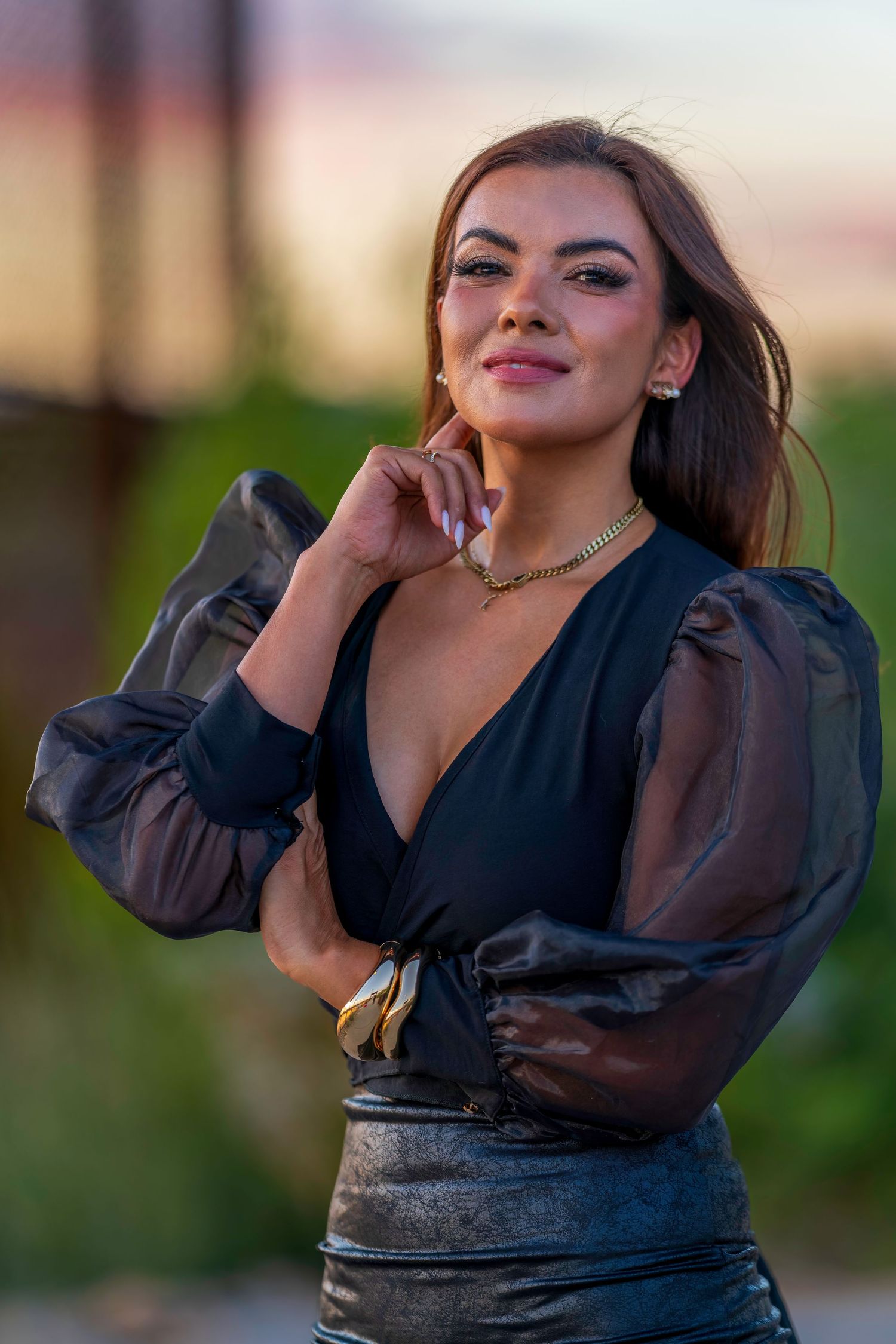 Glamorous woman in black sheer blouse with gold necklace posing at colorful sunset.