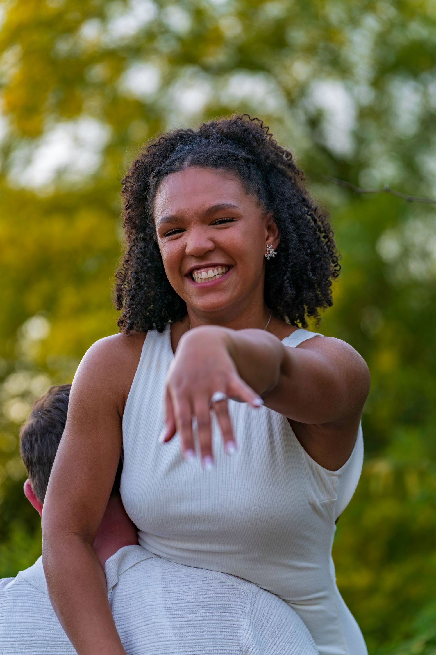 A joyful young woman with curly hair in a white dress laughs outdoors, extending her hand toward the camera.