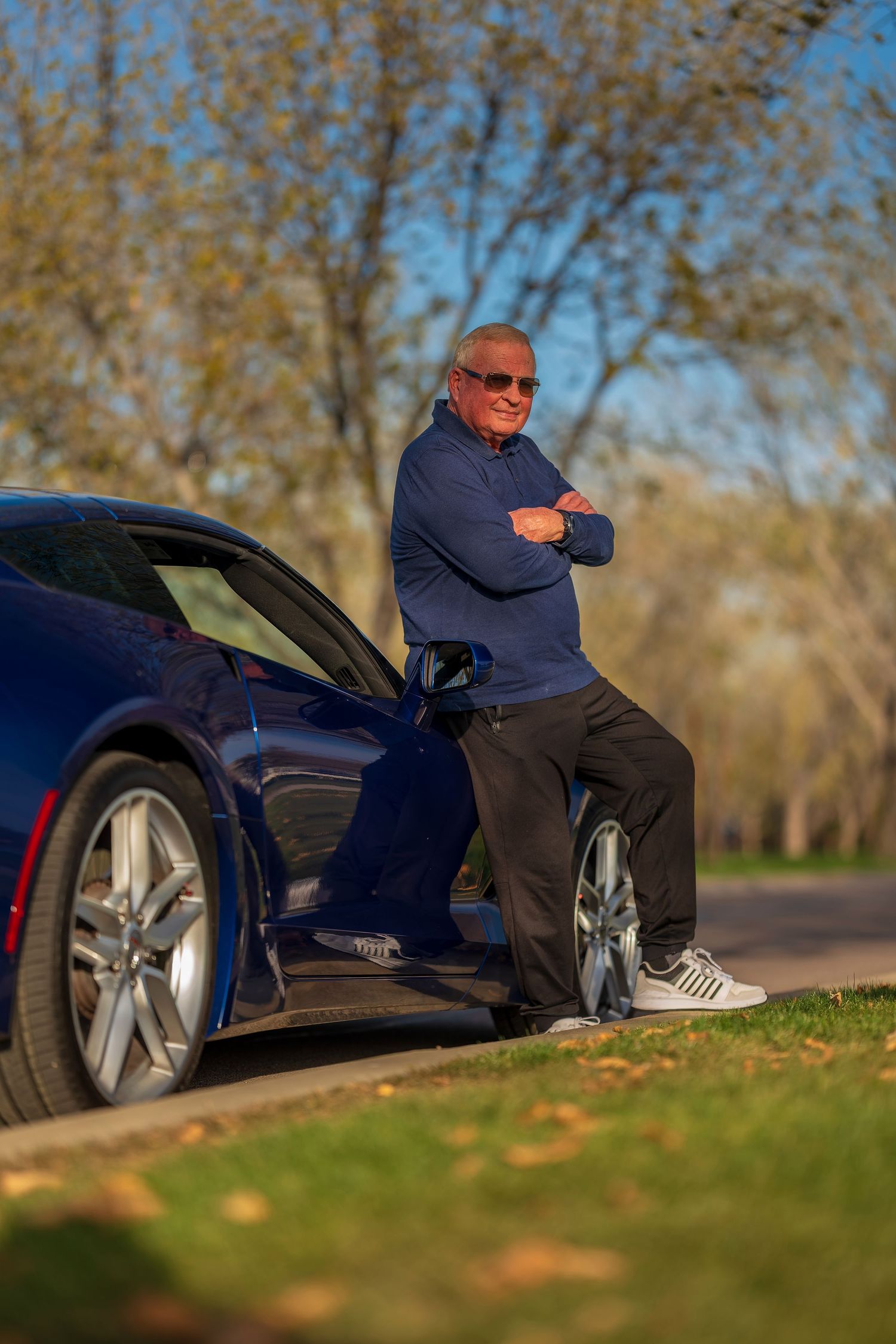 Older man wearing sunglasses leans against blue sports car with arms crossed in sunny park setting.