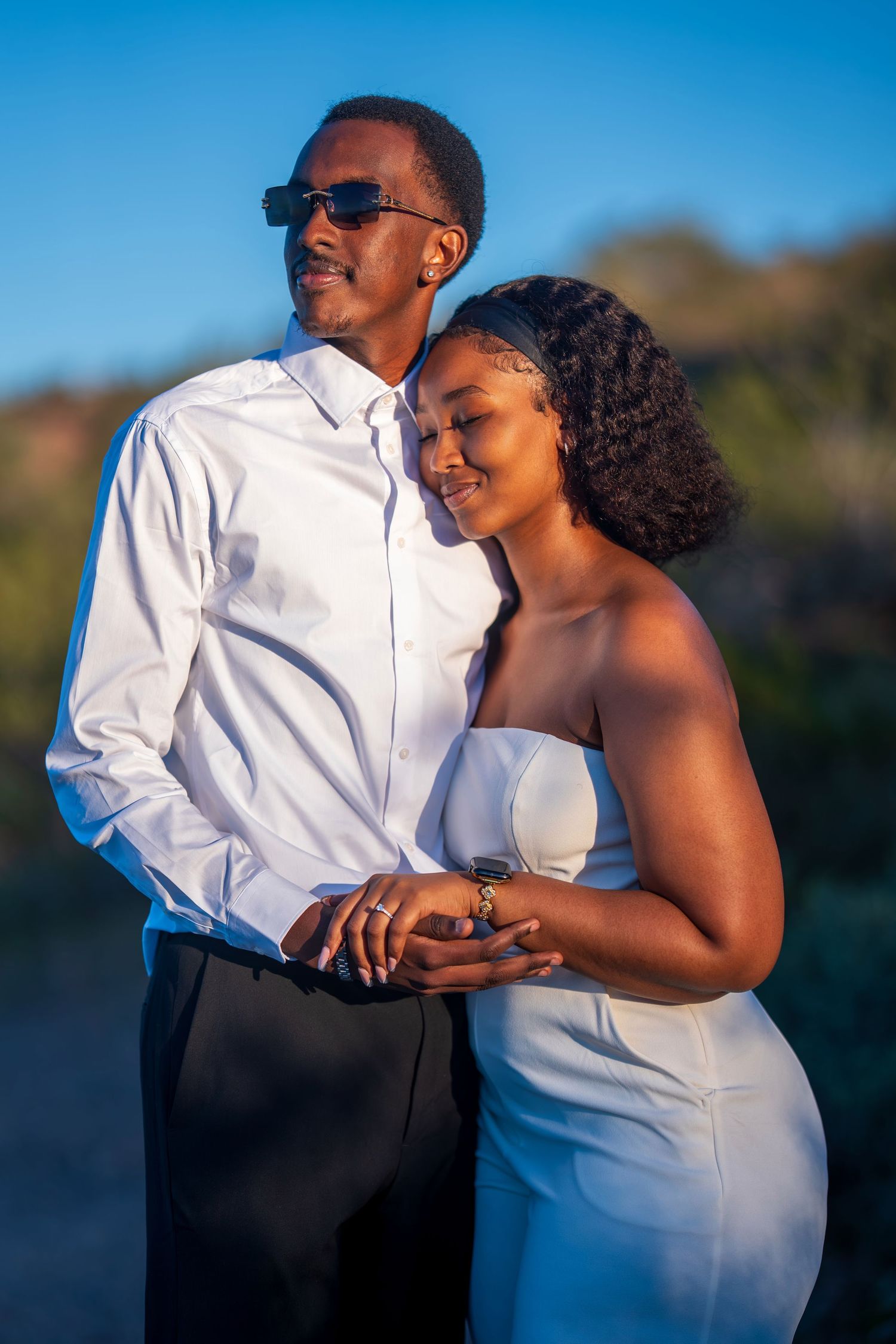 Couple in matching white outfits embracing outdoors at golden hour, man in sunglasses and woman smiling softly.