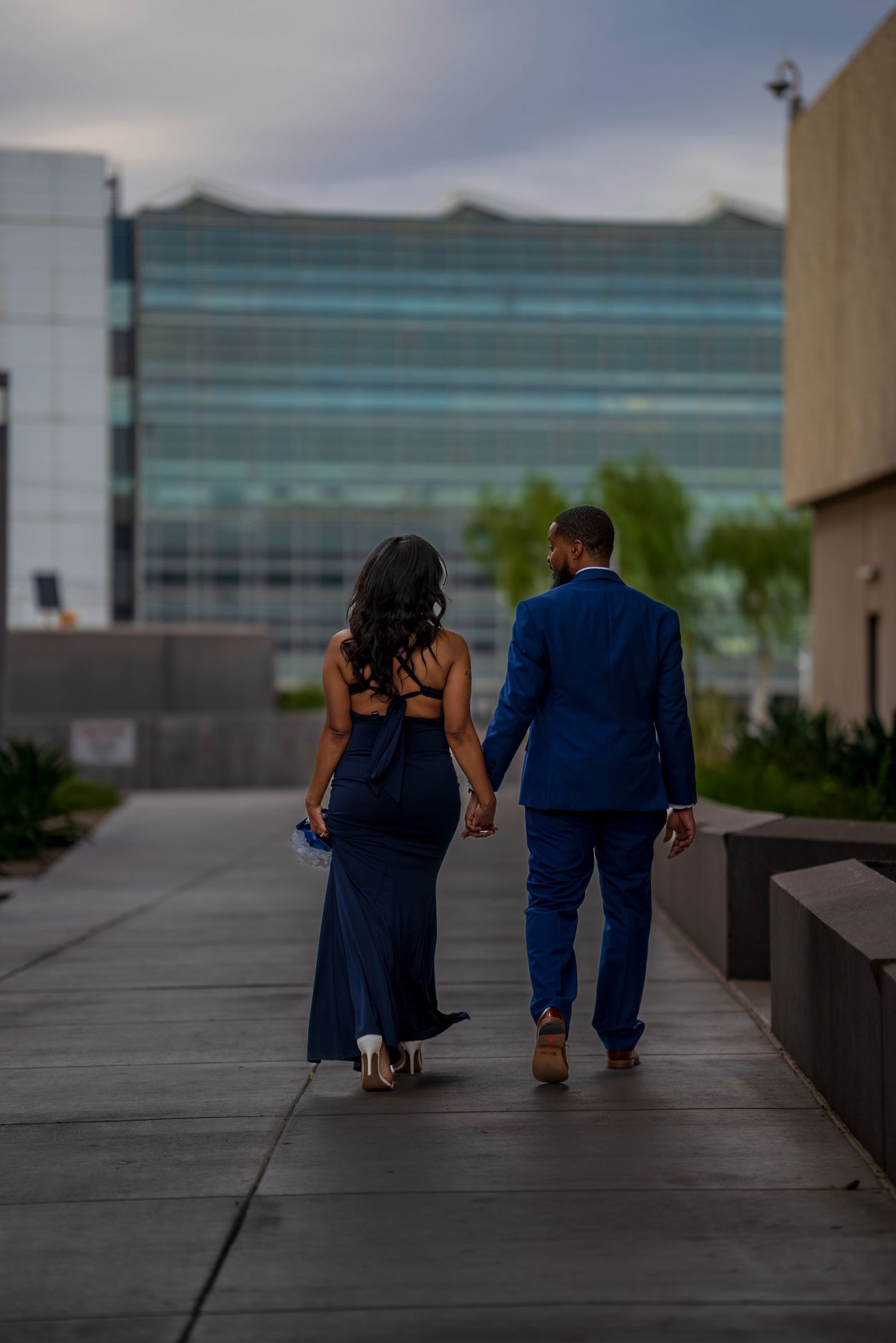 Couple in matching navy blue formal attire walking hand in hand toward modern glass building.
