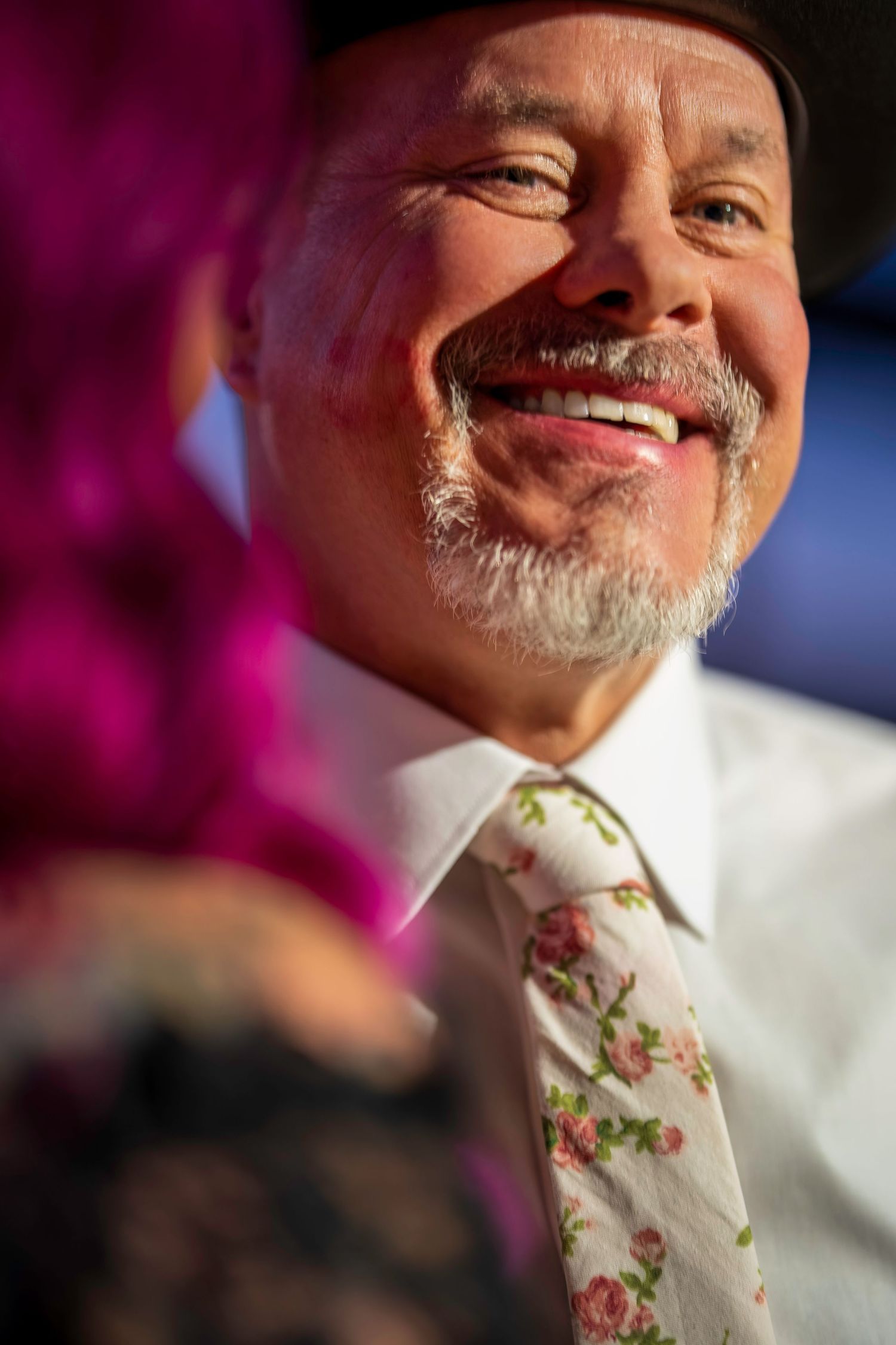 Close-up of smiling older man with gray beard wearing floral tie and gray hat, pink hair visible in foreground.