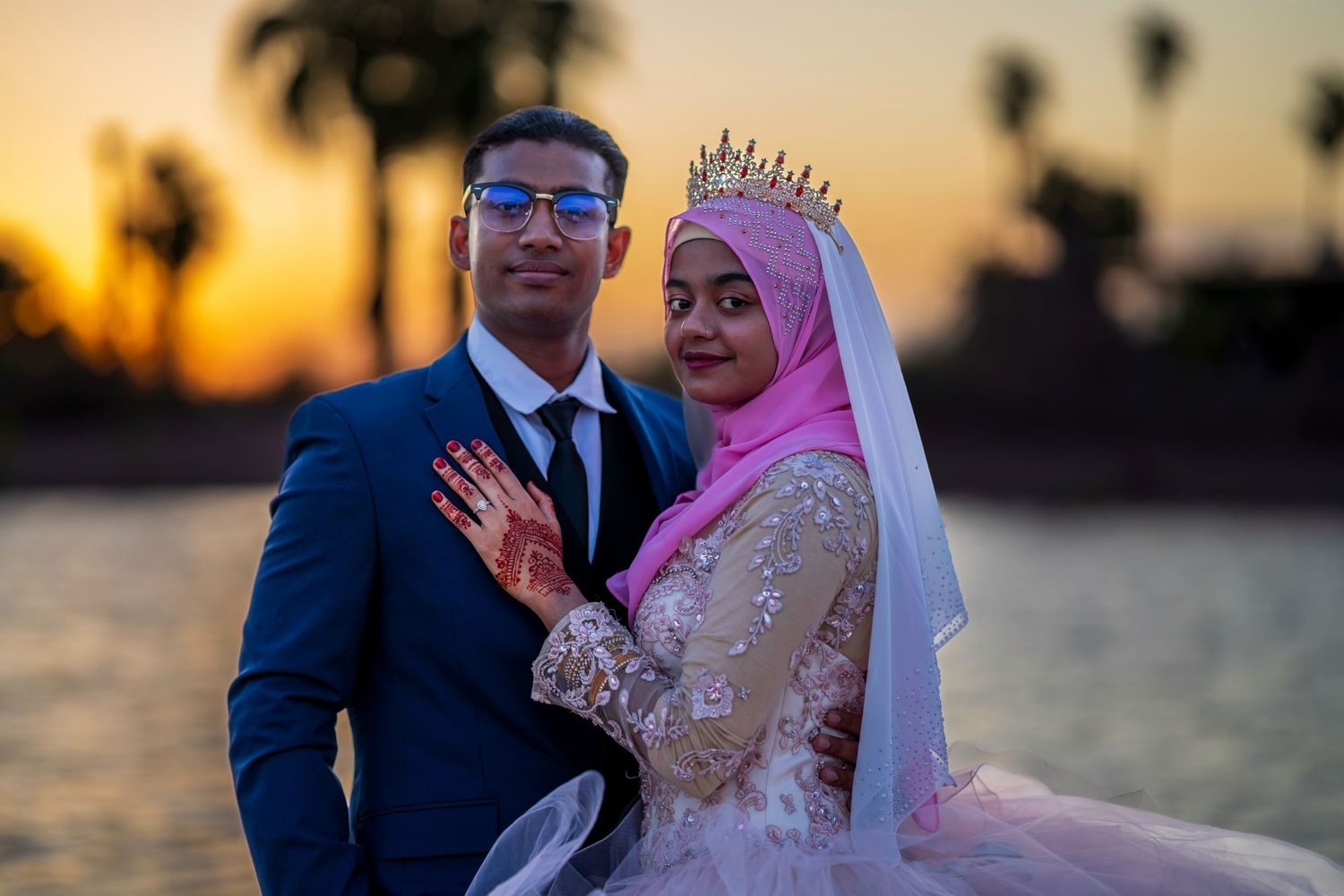 Muslim bride in pink hijab and crown with groom in blue suit posing at sunset waterfront wedding photo.
