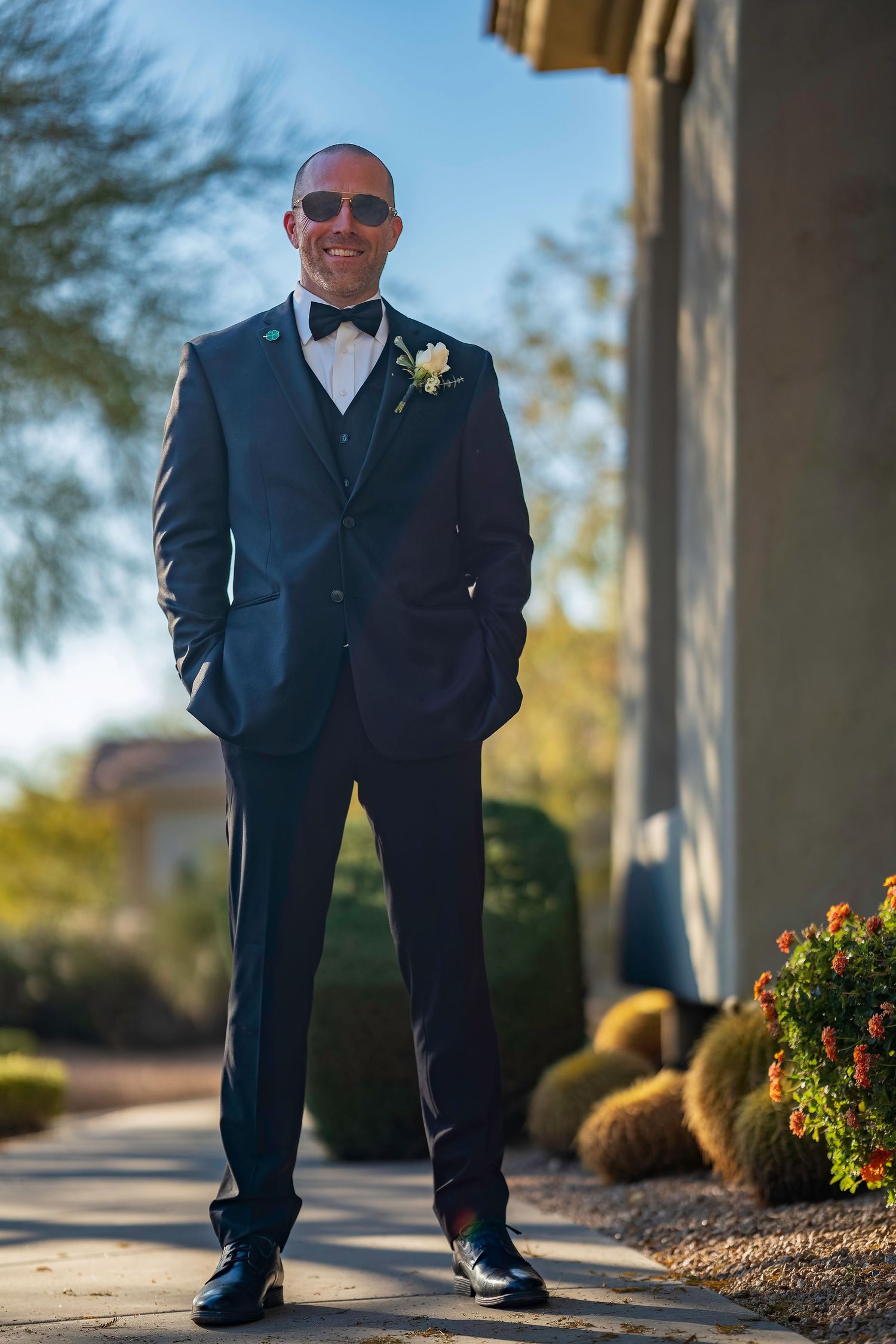 Dapper groom in navy tuxedo with bow tie and boutonniere posing outdoors in sunglasses on wedding day.