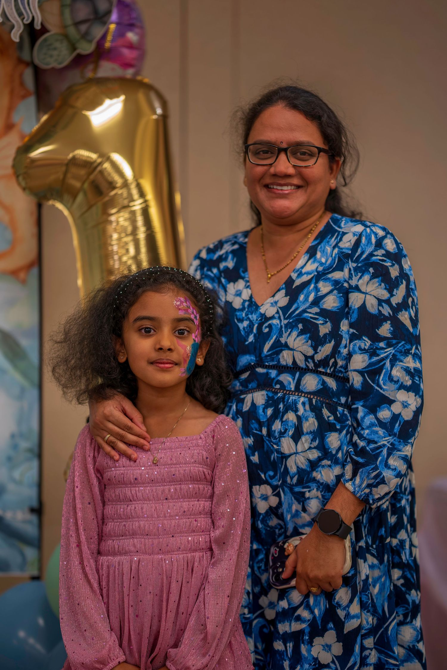 Young girl with face paint poses with smiling woman in blue floral dress at party.