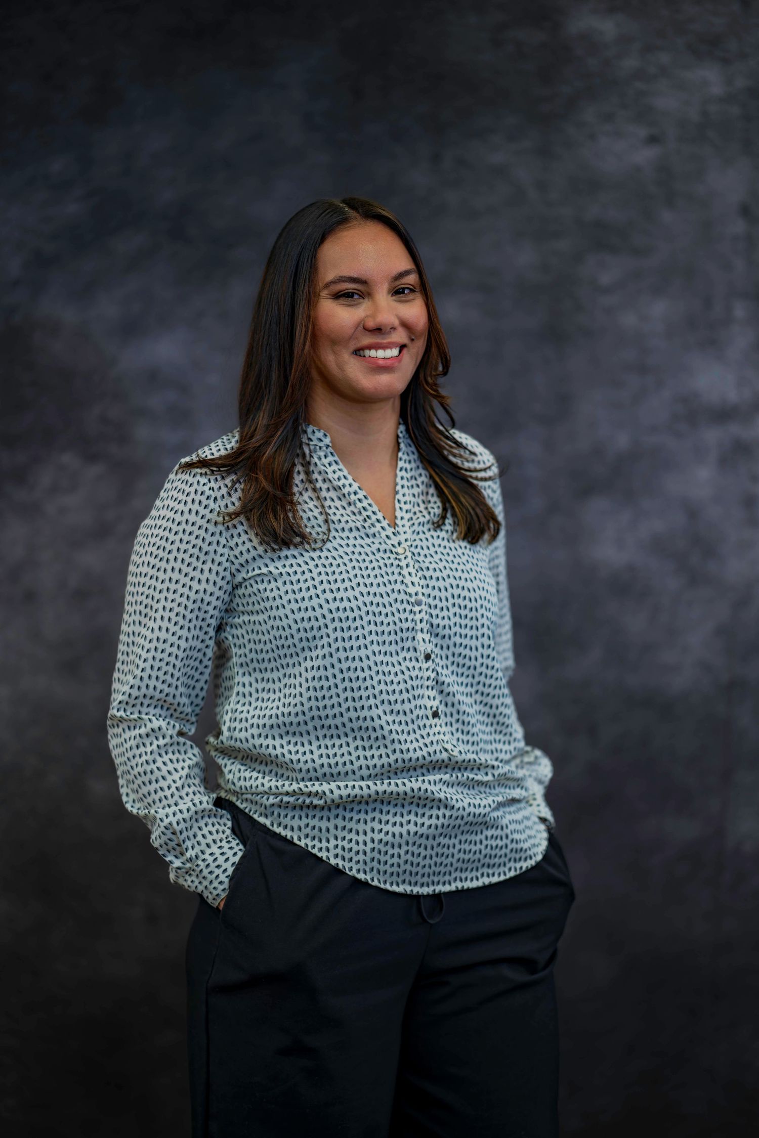 Professional portrait of person wearing patterned button-up blouse against dark background.