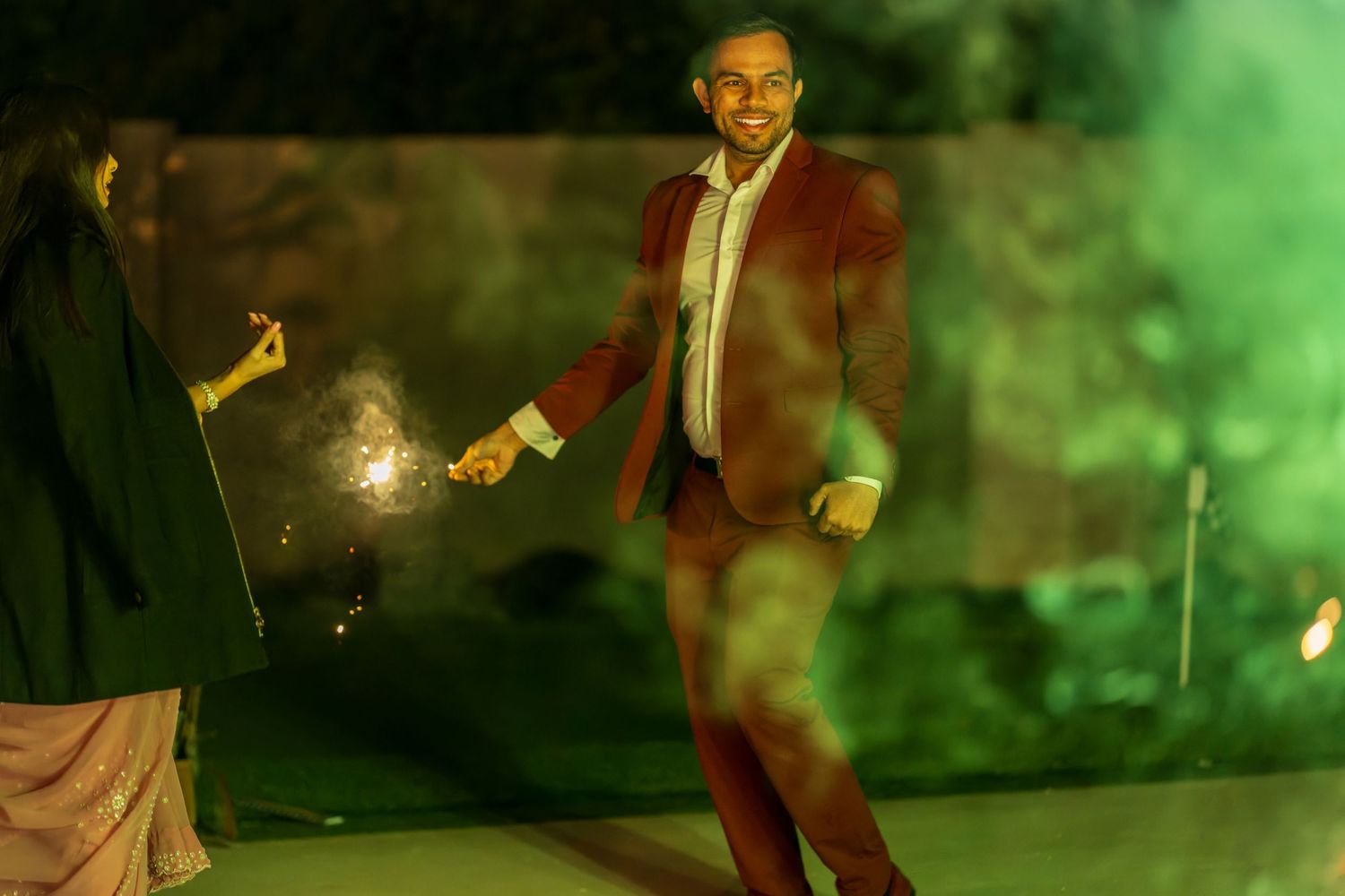 A person in a burgundy suit stands near sparklers in atmospheric green lighting.