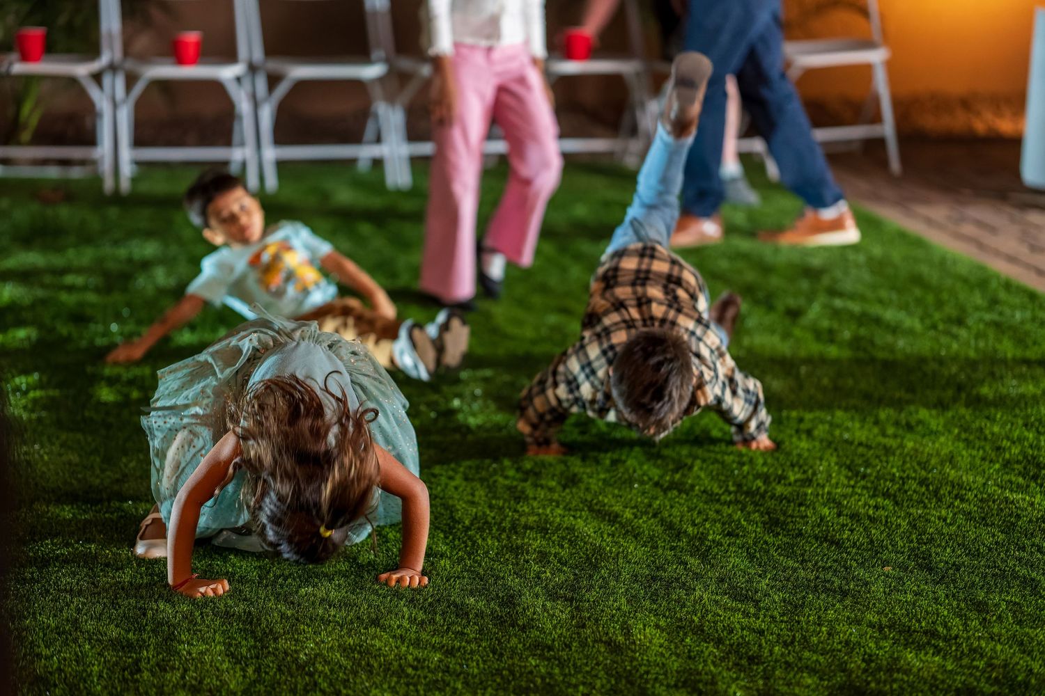 Children playing and crawling on grass near folding chairs at an outdoor event.