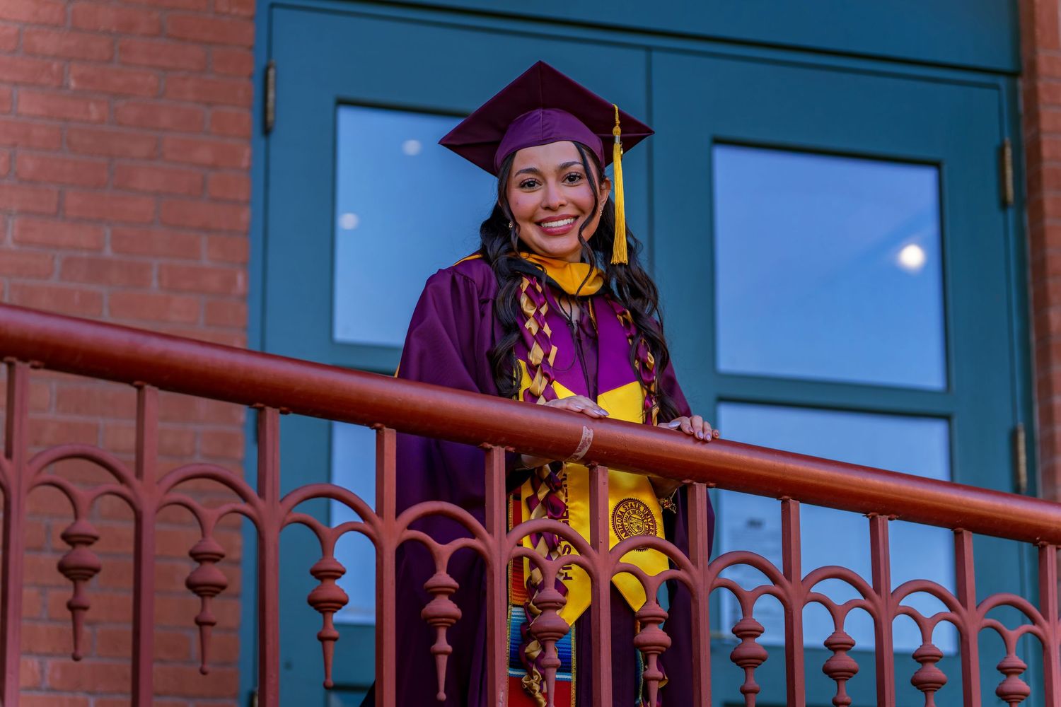 Graduate in academic regalia leans on ornate railing during sunset graduation photoshoot.