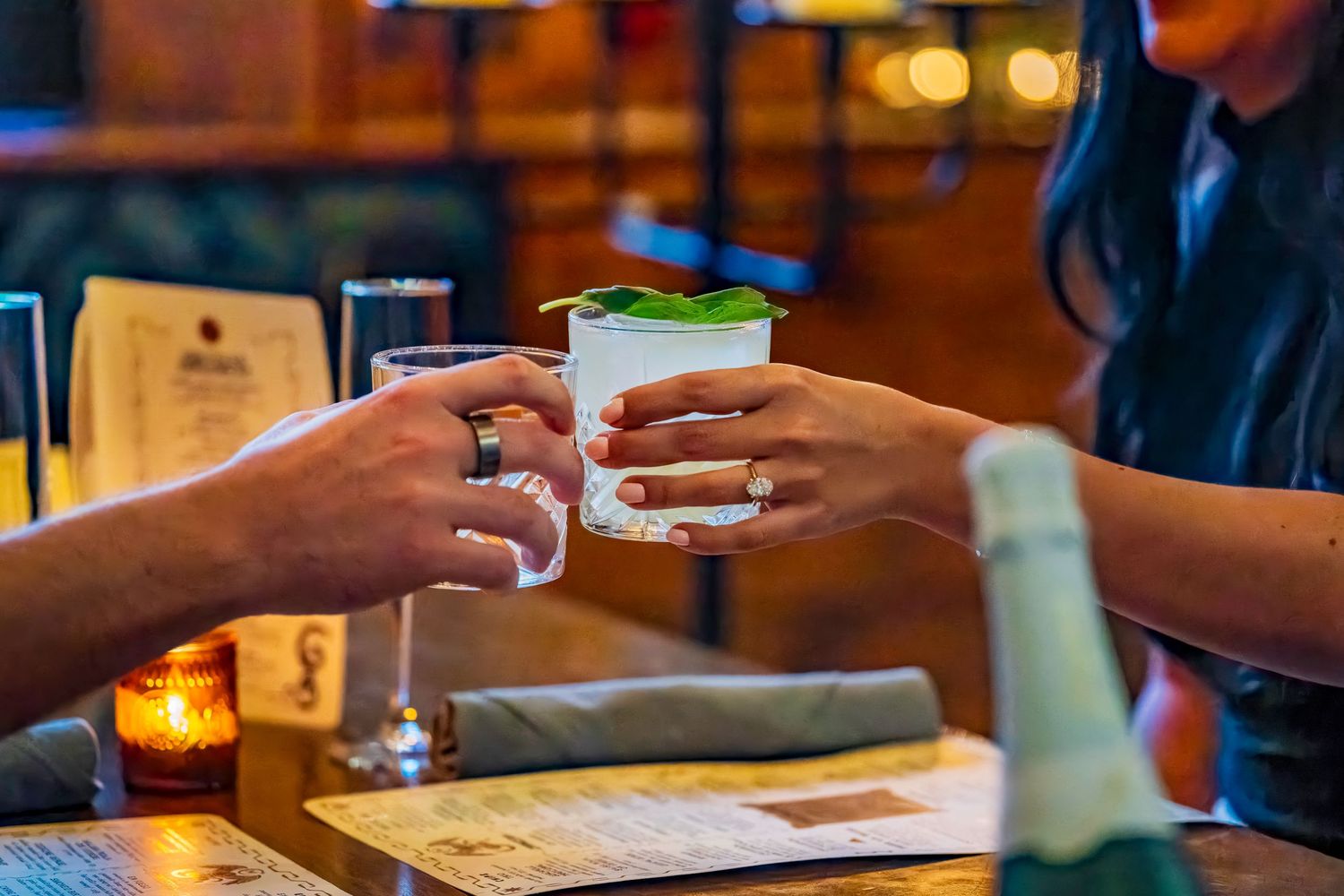 Two hands toast with cocktail glasses in a warmly lit bar setting.