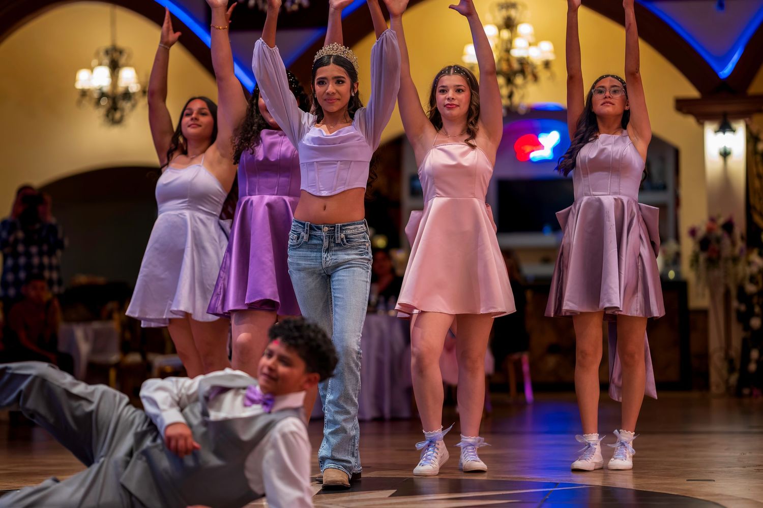 Young women in matching purple and pink outfits perform a choreographed dance routine at an elegant indoor celebration.