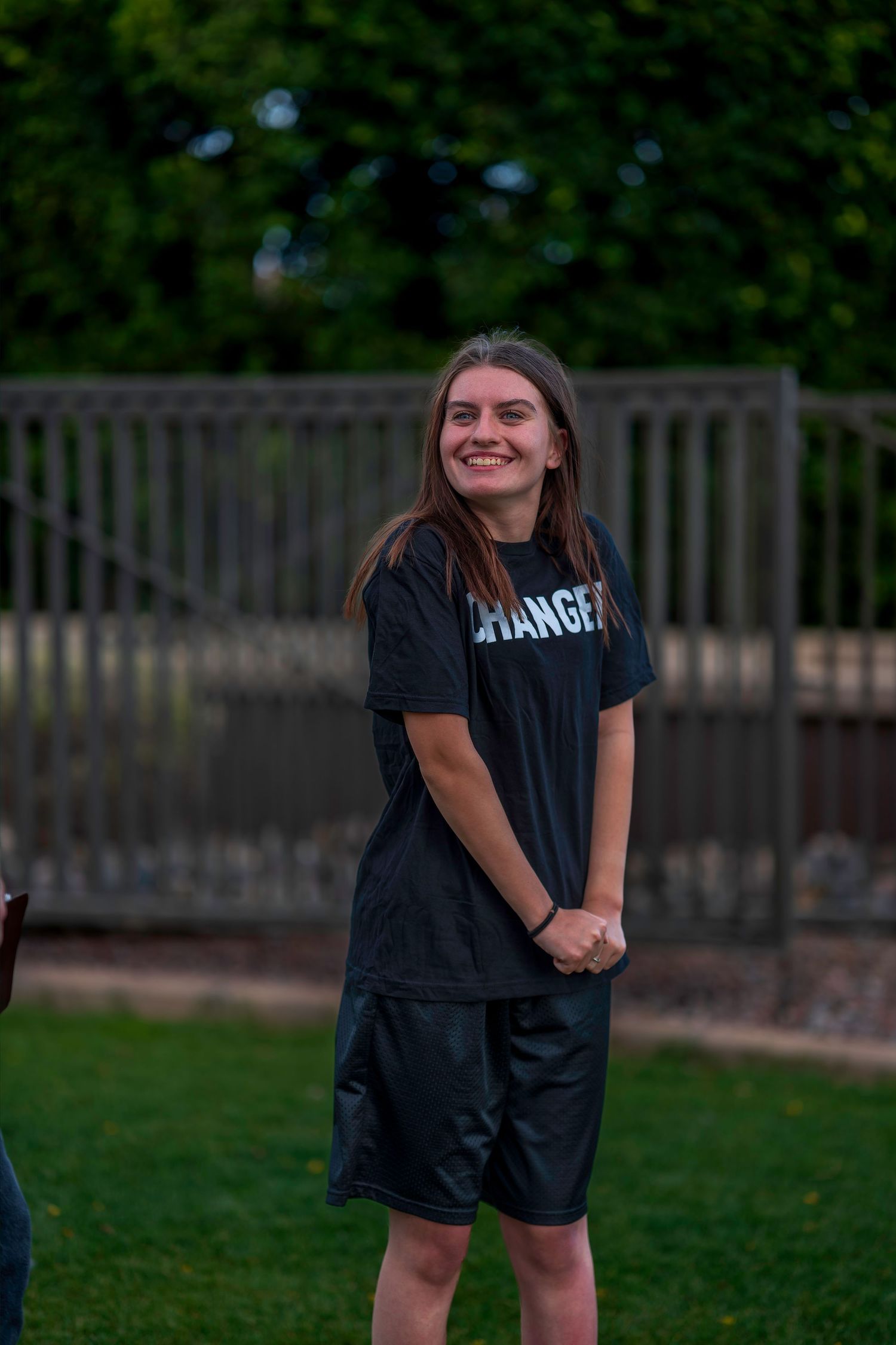 Smiling young woman in black Rangers t-shirt and shorts standing on green grass outdoors.