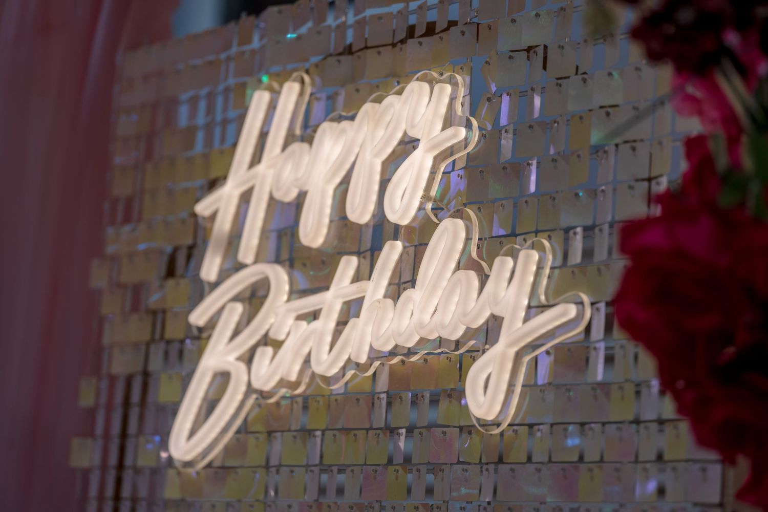 Glowing neon Happy Birthday sign on a shimmering gold sequin wall backdrop with pink floral decor.