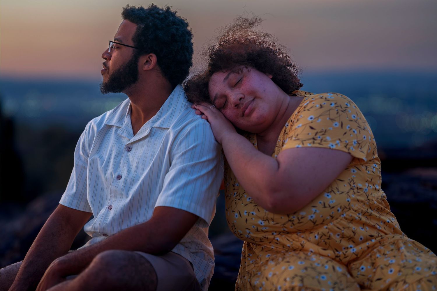 Couple sits together at sunset, woman in yellow dress resting her head on mans shoulder romantically.