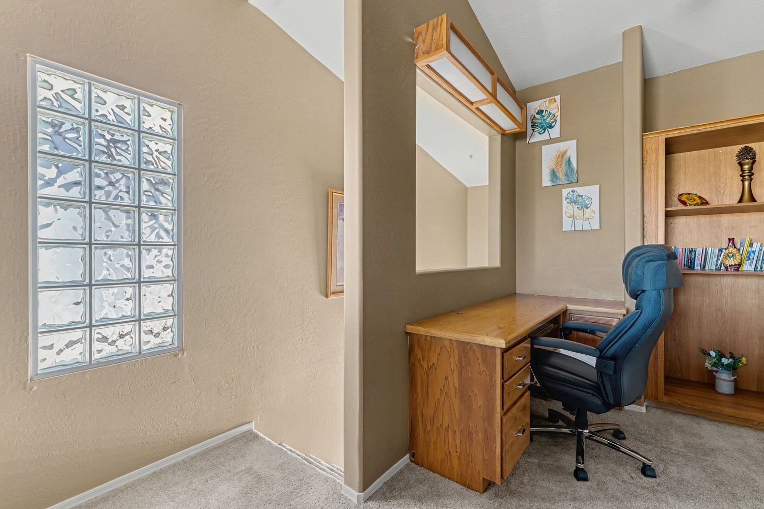 Home office nook with wooden desk, blue chair, glass block window, and built-in shelving unit.