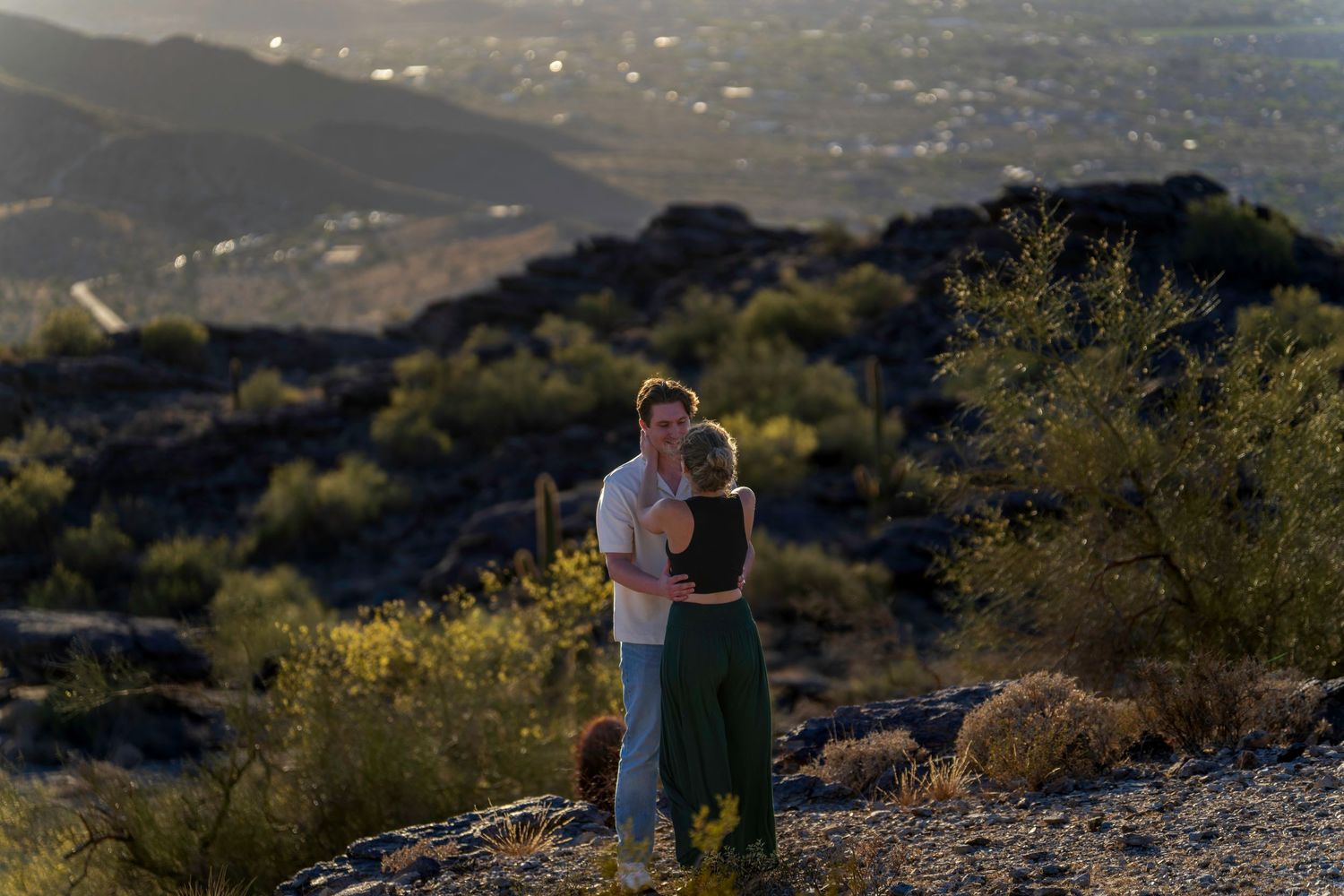 Couple embracing at sunset on a rocky desert overlook with mountains and city in the background.