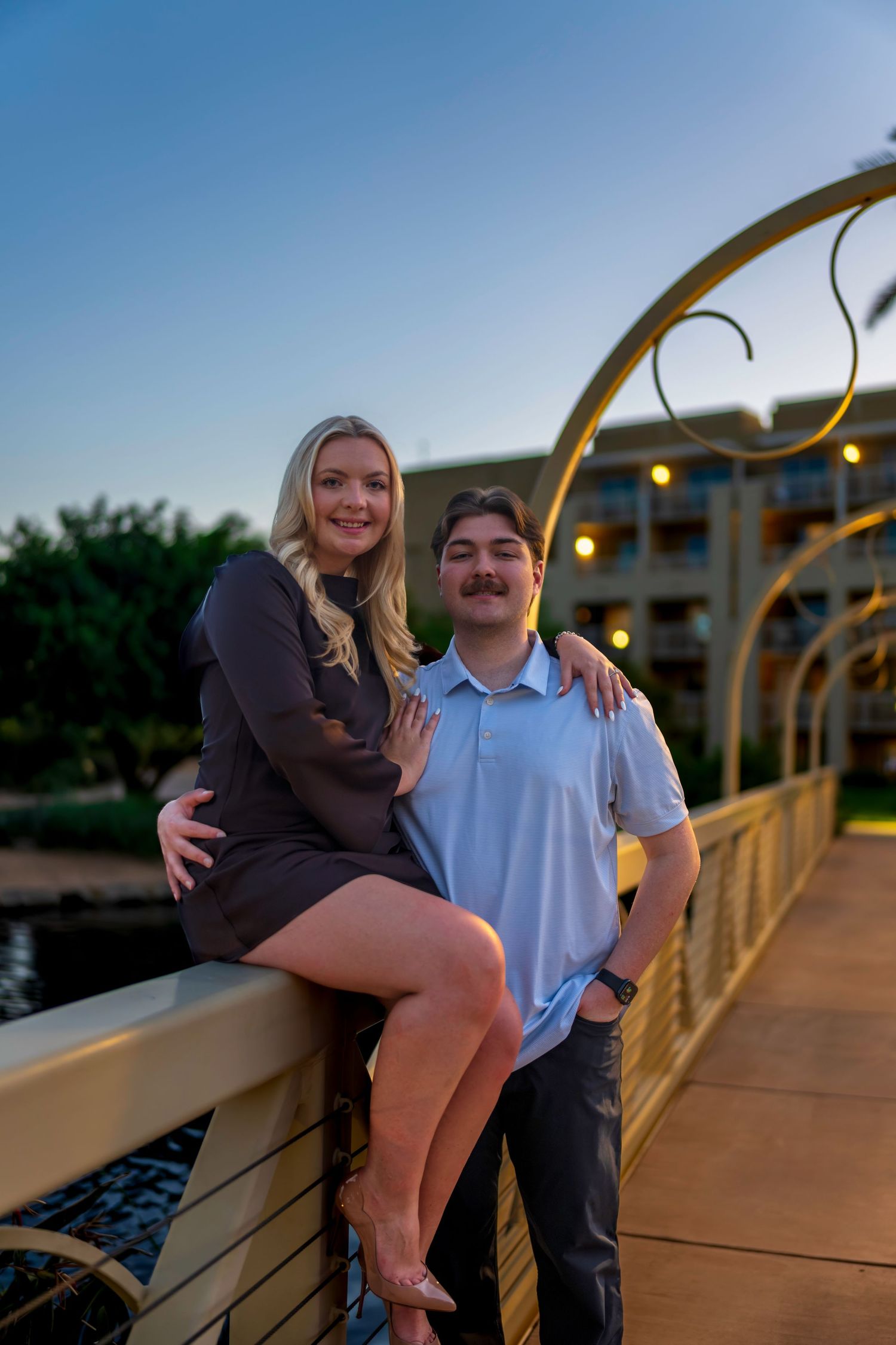 Smiling engaged couple posing on a decorative golden bridge at dusk near a resort waterway.