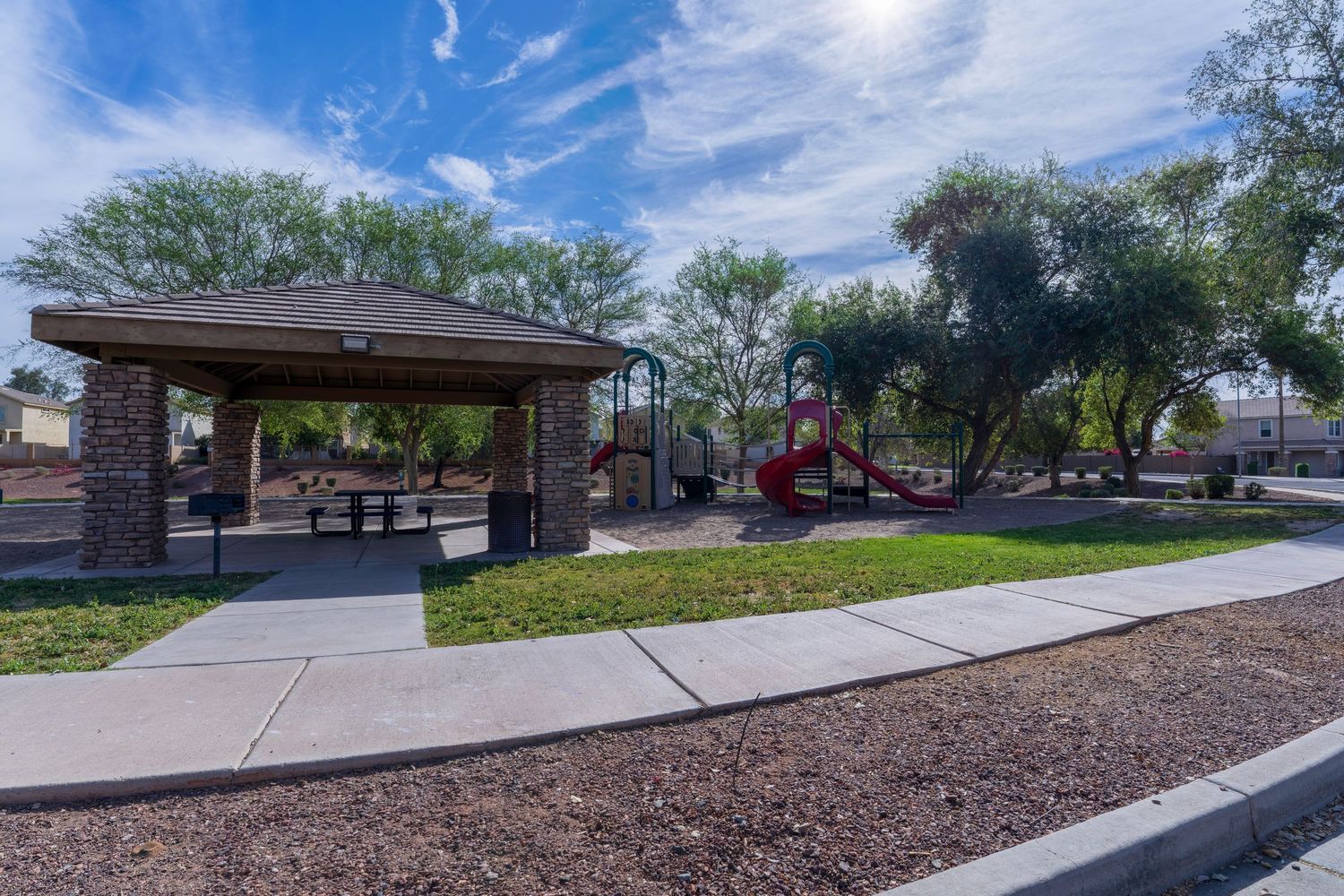 Community park featuring a covered picnic pavilion, red playground equipment, and lush trees on a sunny day.