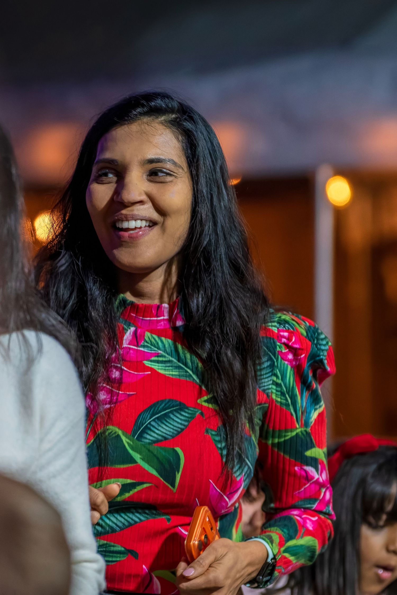 A person wearing a vibrant red and green tropical print top smiles warmly in an indoor setting.
