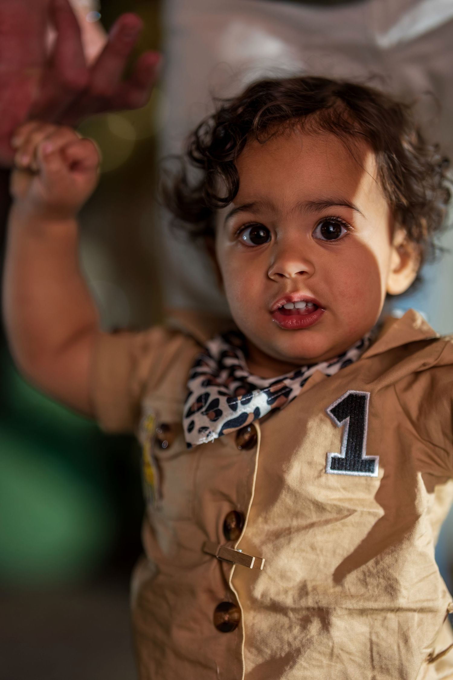 A toddler wearing a tan outfit with a number 1 makes a peace sign while smiling at the camera.