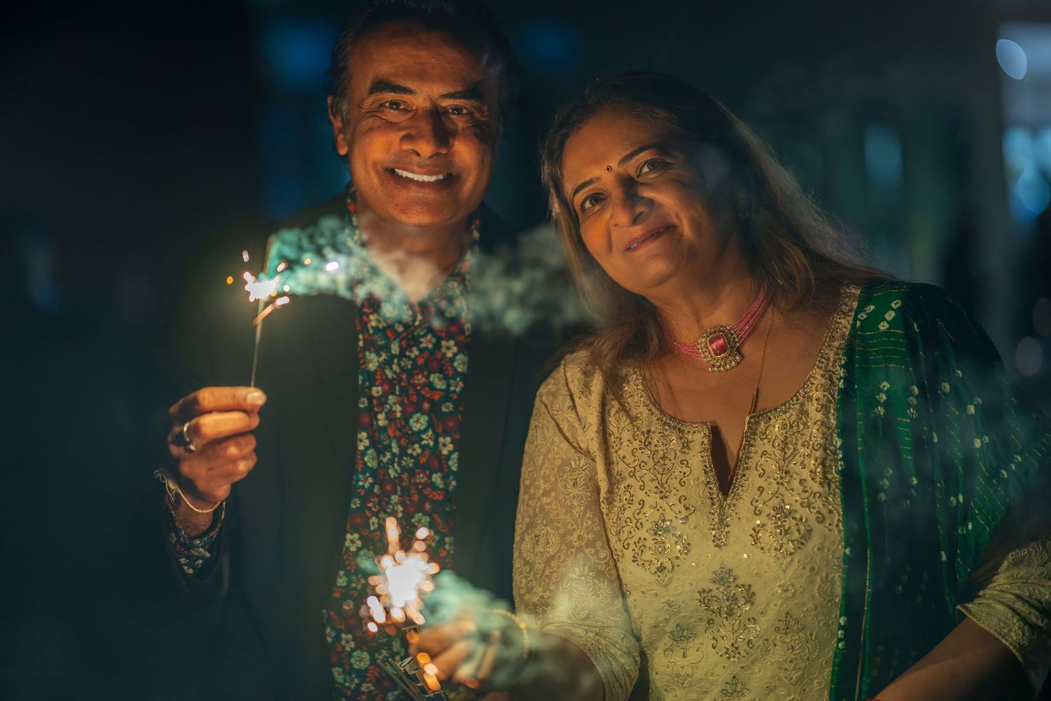Two people in formal wear smile while holding sparklers during an evening celebration.
